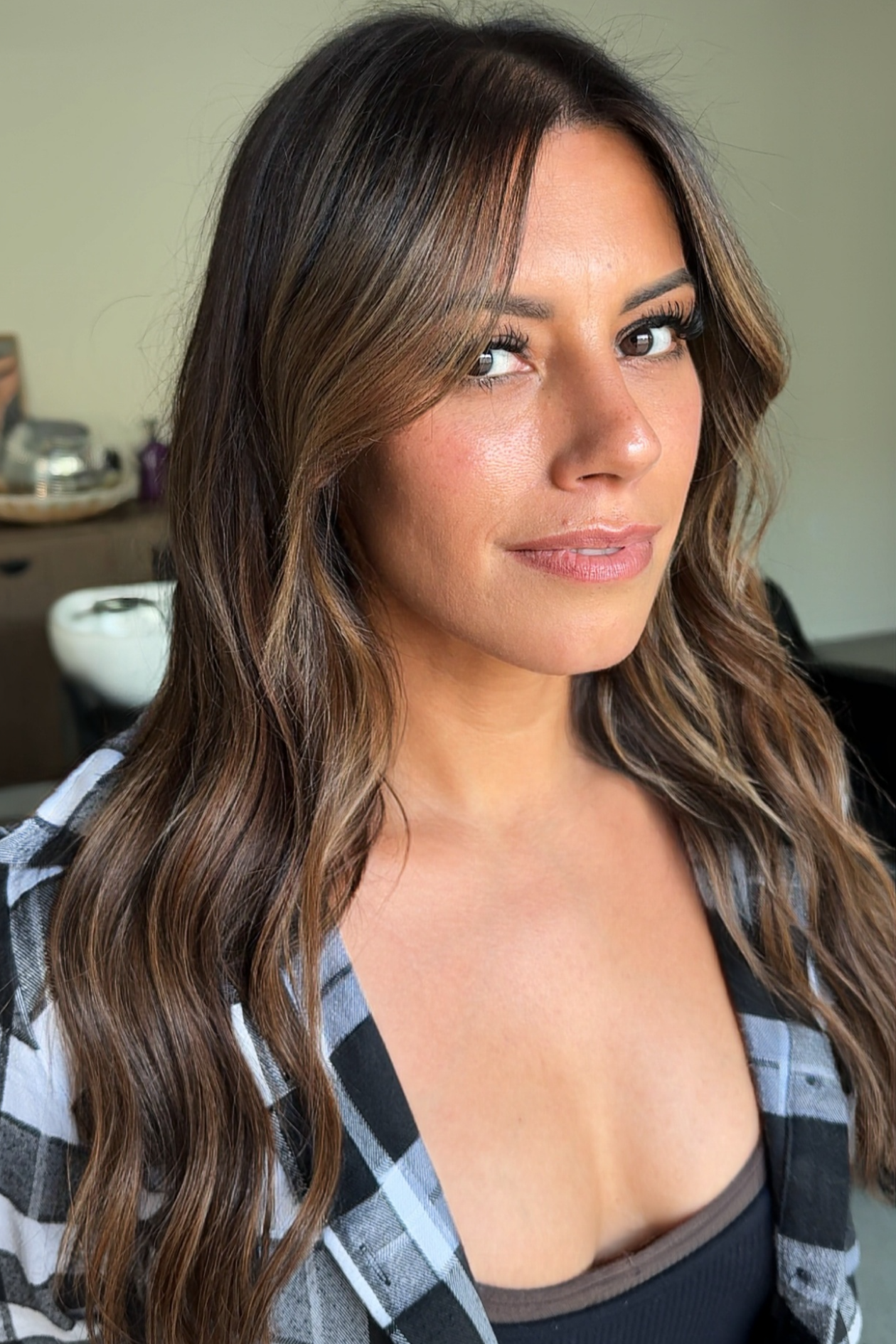 A woman with long, wavy brown hair and makeup, wearing a black top under a black and white plaid shirt, standing indoors with a neutral background.
