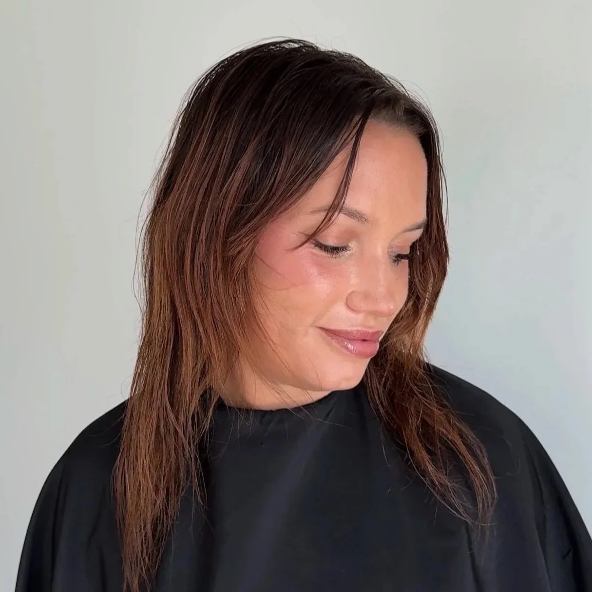 A woman with shoulder-length brown hair wearing a black salon cape looking down and smiling against a plain light background.