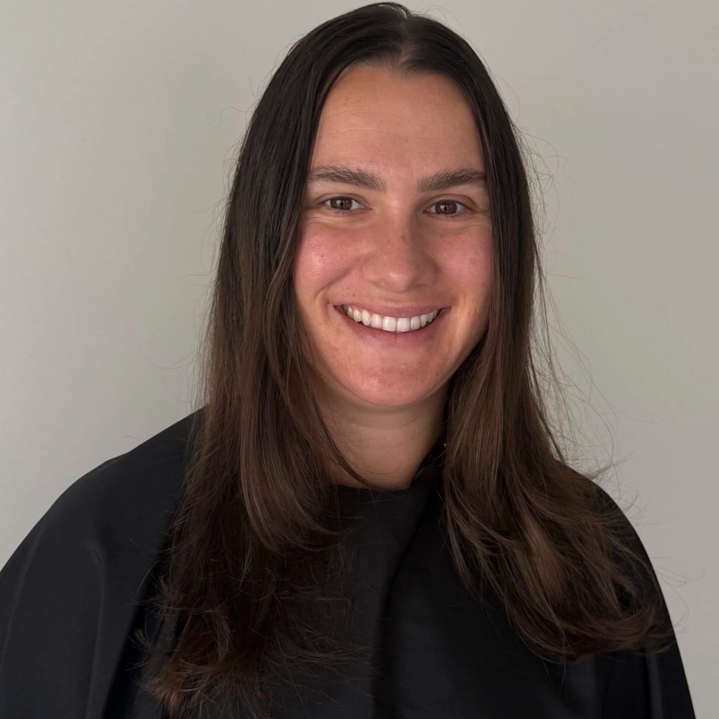 Woman with long brown hair smiling, wearing a black haircut cape, standing against a plain light gray wall.