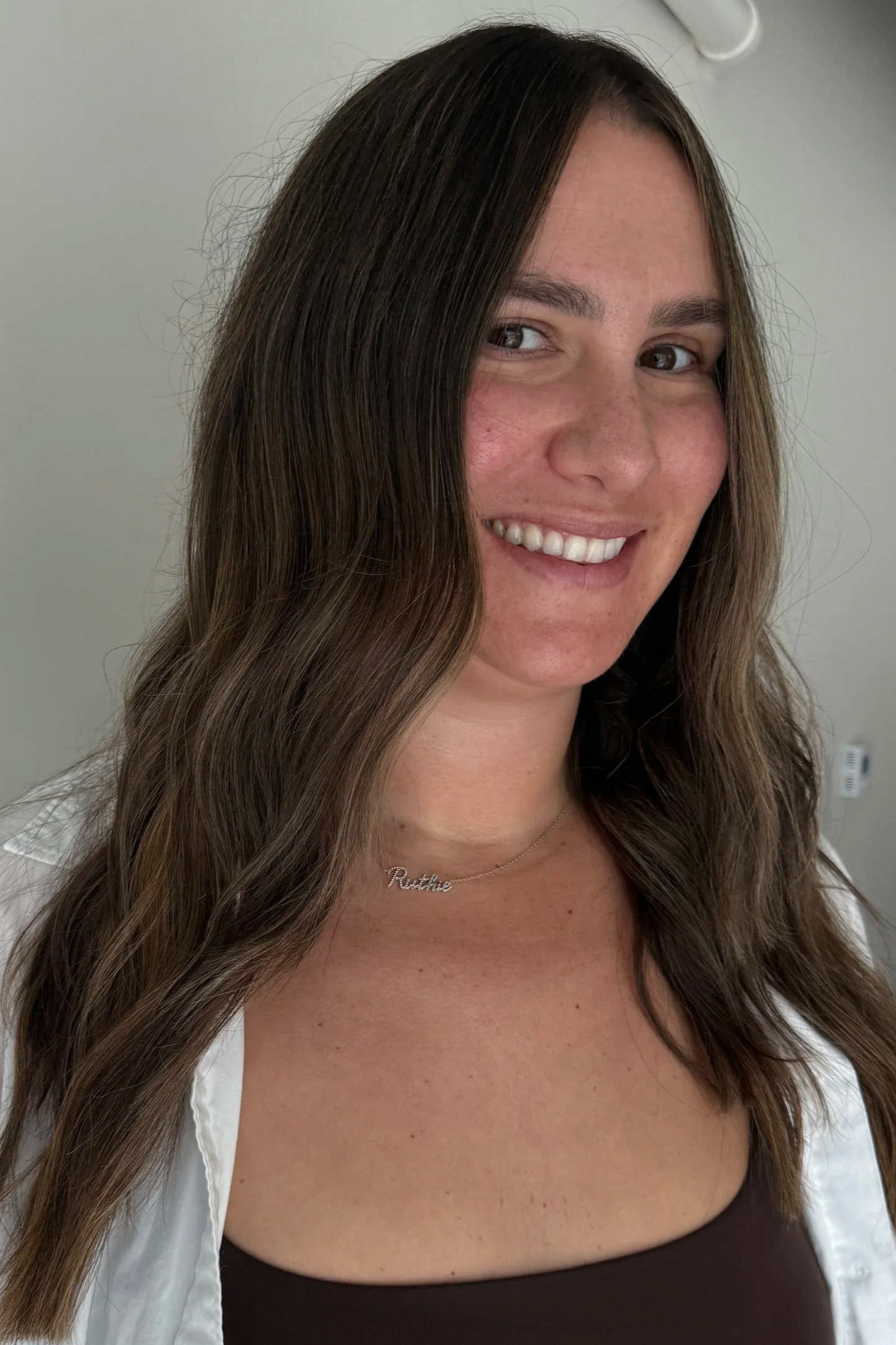 A woman with long wavy brown hair, smiling, wearing a white open shirt over a dark brown top, and a necklace with the name 'Ruthie' in cursive, standing indoors against a plain wall.