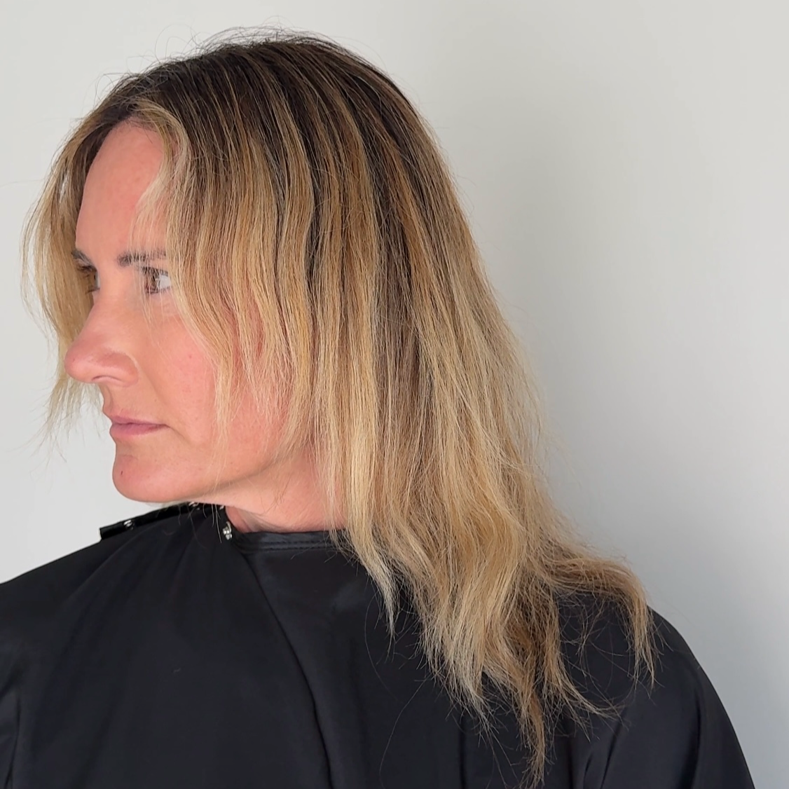 A woman with medium-length, wavy blonde hair with darker roots, wearing a black salon cape, looking to the left against a plain white background.