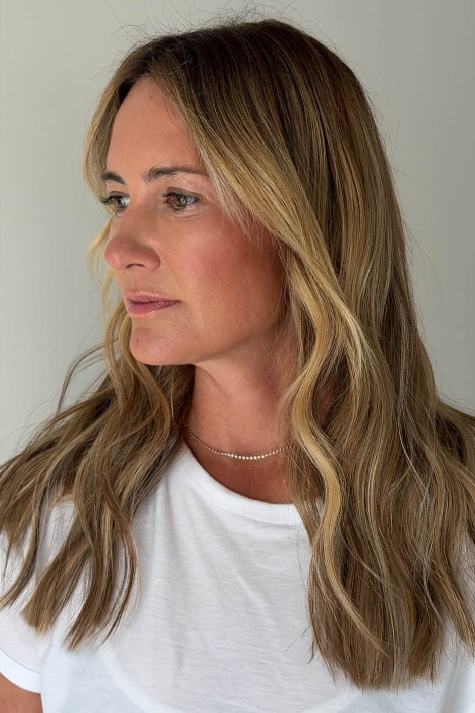 A woman with long, wavy, light brown hair with blonde highlights wearing a white t-shirt and a delicate silver necklace, looking to her left against a neutral background.
