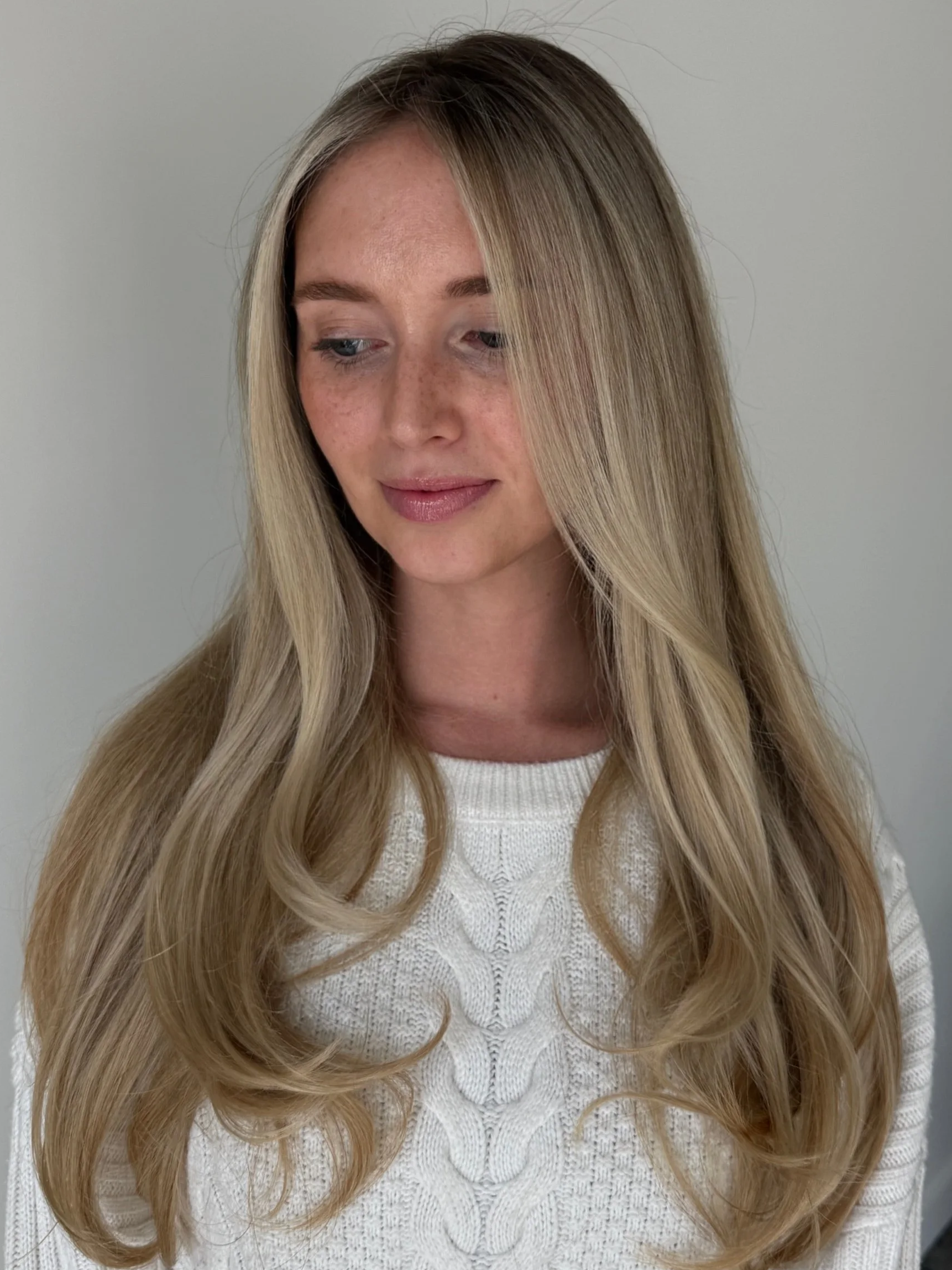 A young woman with long, wavy blonde hair, wearing a white cable-knit sweater, looking down with a gentle expression, standing against a plain white wall.