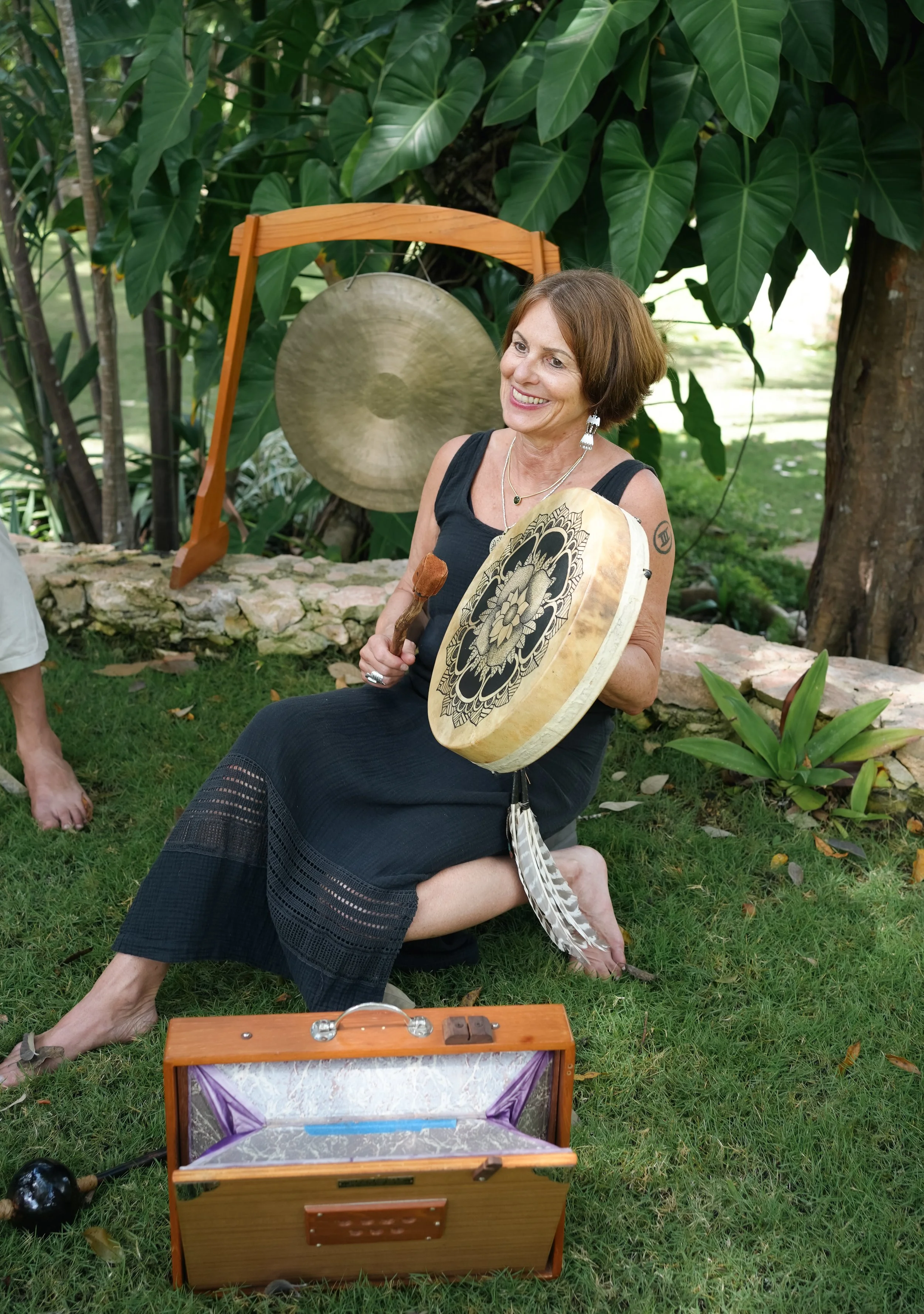 A woman with short brown hair sitting outdoors on grass, holding a decorated drum and a mallet, smiling. She is dressed in a black dress with a tattoo on her upper arm. Behind her are large green leaves, a large gong, and a small wooden box with a handle, open to reveal a soft interior.