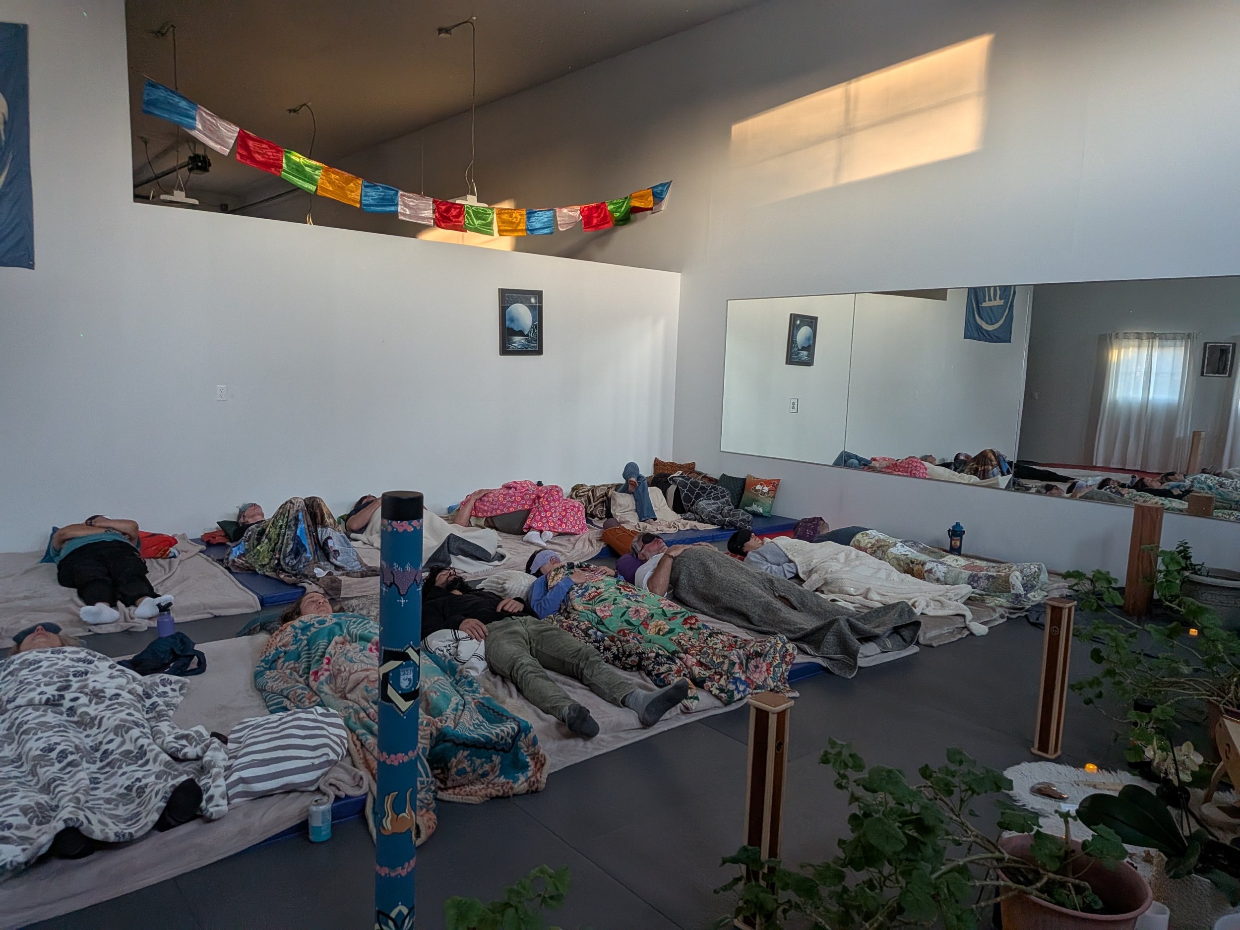 Multiple adults lying on mats on the floor in a room with a large mirror, a flag, and a colorful banner hanging from the ceiling, possibly resting or sleeping.