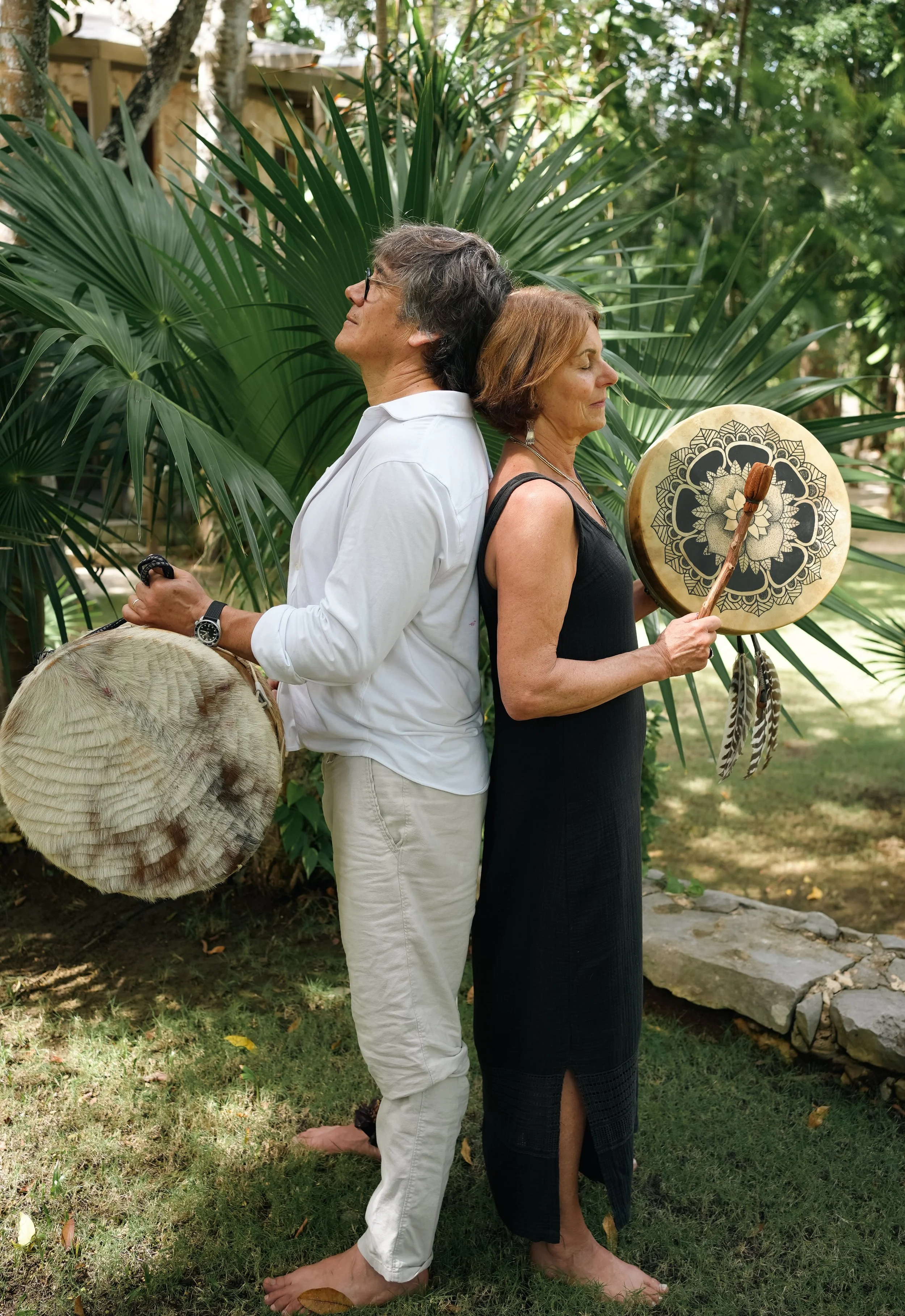 A man and a woman stand back to back outdoors in a garden with lush green foliage, both barefoot with eyes closed. The woman holds a decorated drum with feathers, and the man has a large native drum.