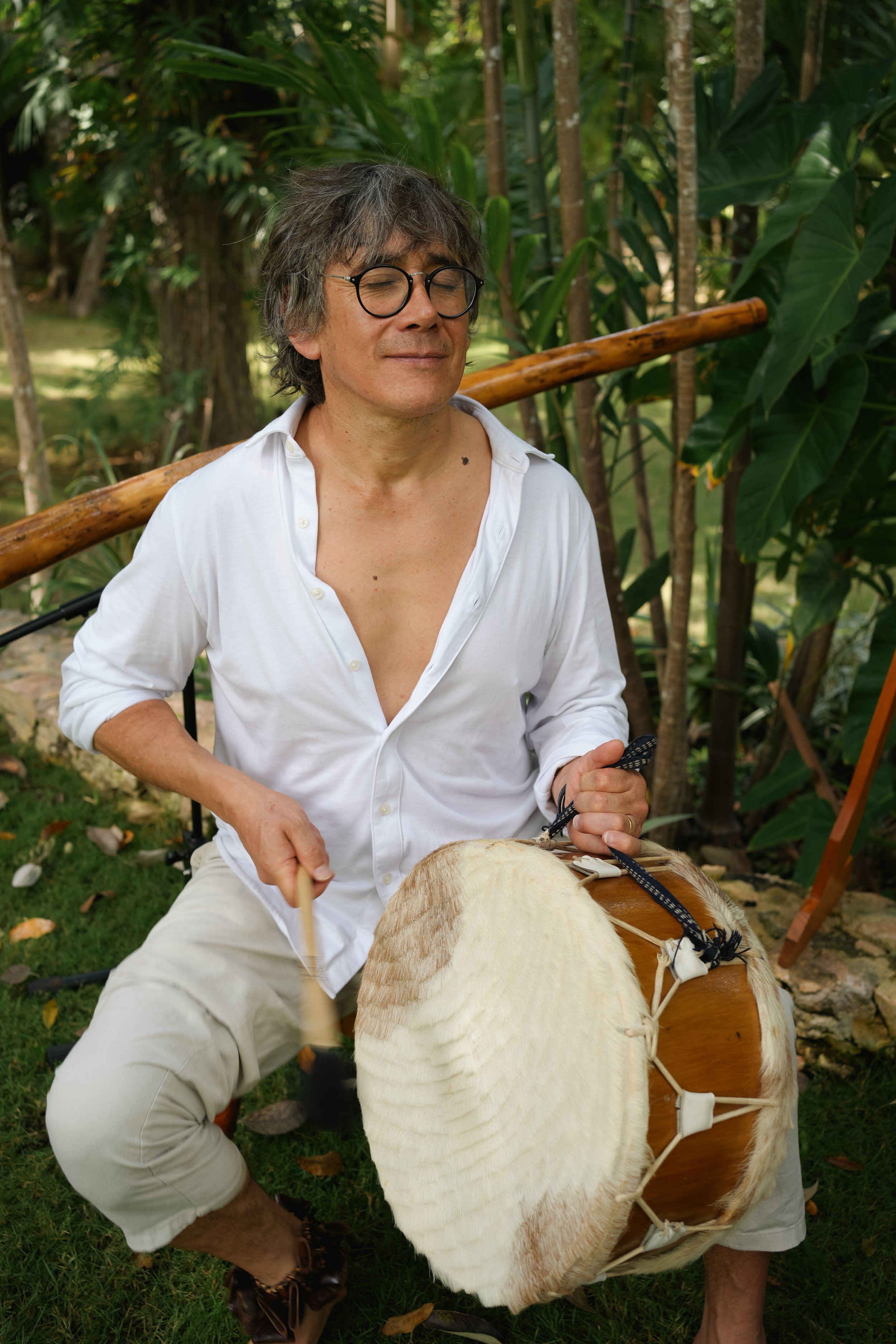 A man with gray hair, glasses, and a white shirt sitting outdoors, holding a drum, surrounded by lush green foliage.