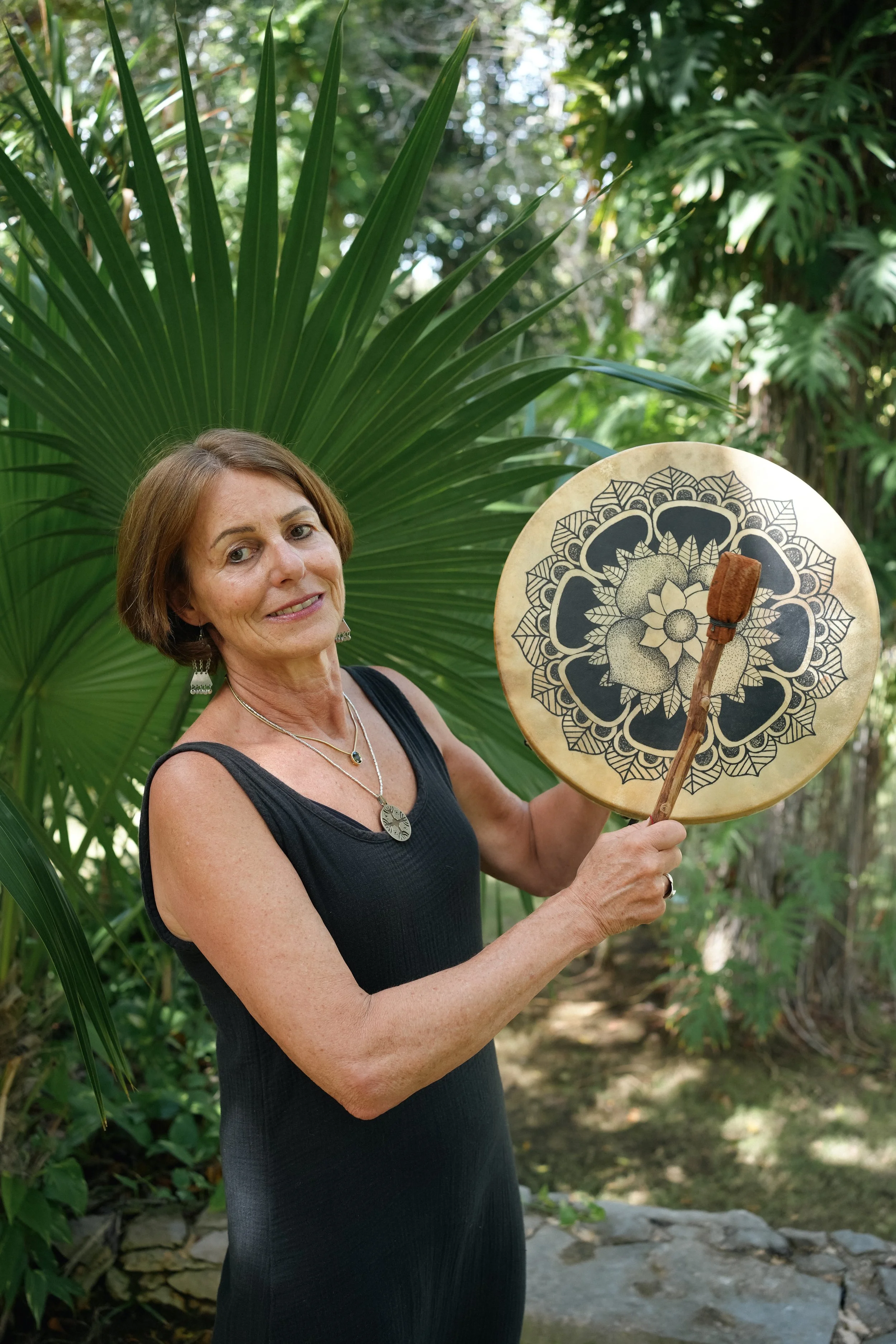 A woman with short brown hair and earrings is standing outdoors among lush green tropical plants, holding a decorative hand drum with a floral design and a small mallet.