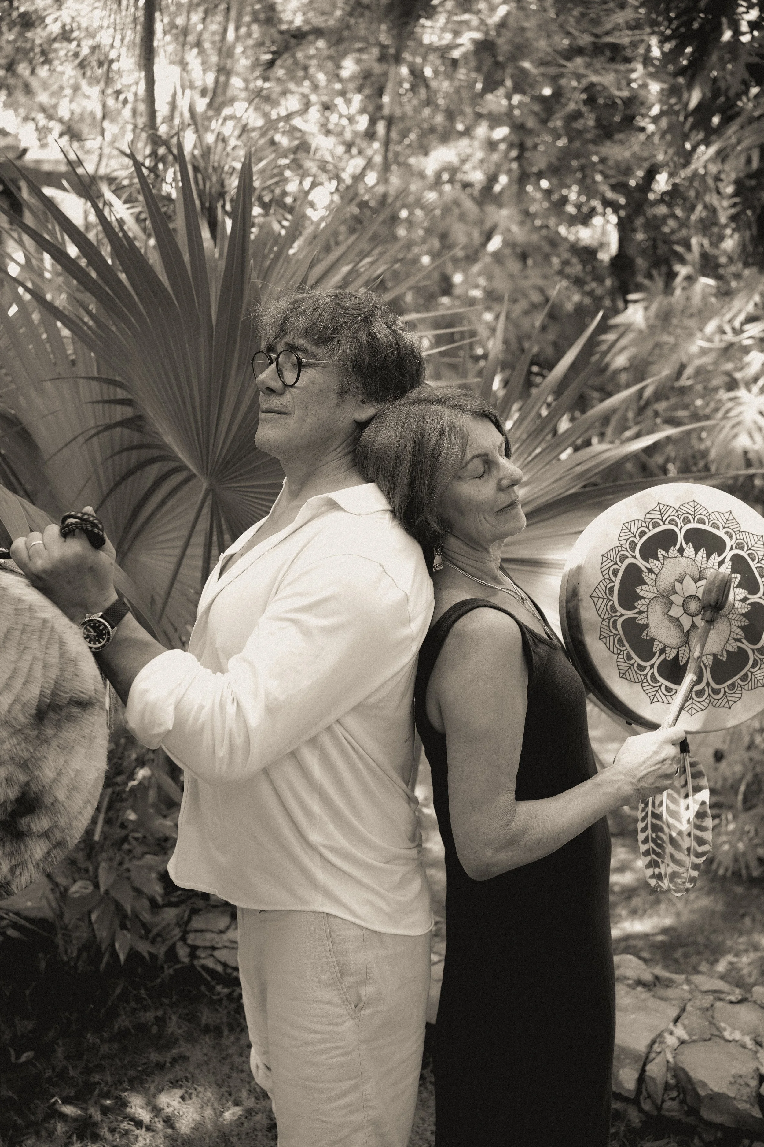 Two people, a man and a woman, standing back to back outdoors, holding fans and surrounded by lush plants and trees.