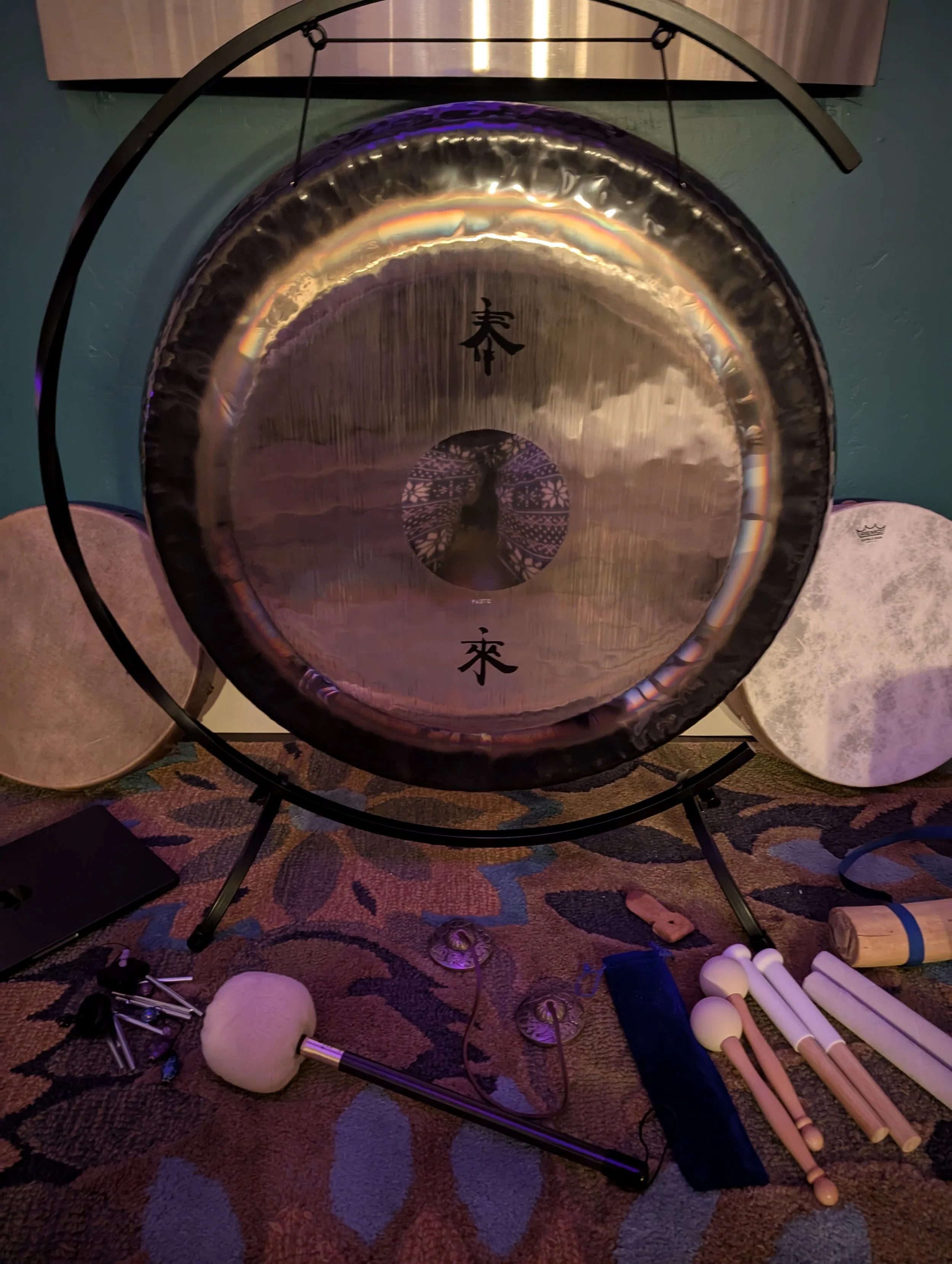 A large gong with Asian characters painted on its surface is the main focus in this room. There are various percussion mallets and beaters scattered around the floor, along with some electronic equipment and a pair of gloves. The room has a patterned carpet and a wall with a teal color, with a musical instrument or object mounted on the wall.