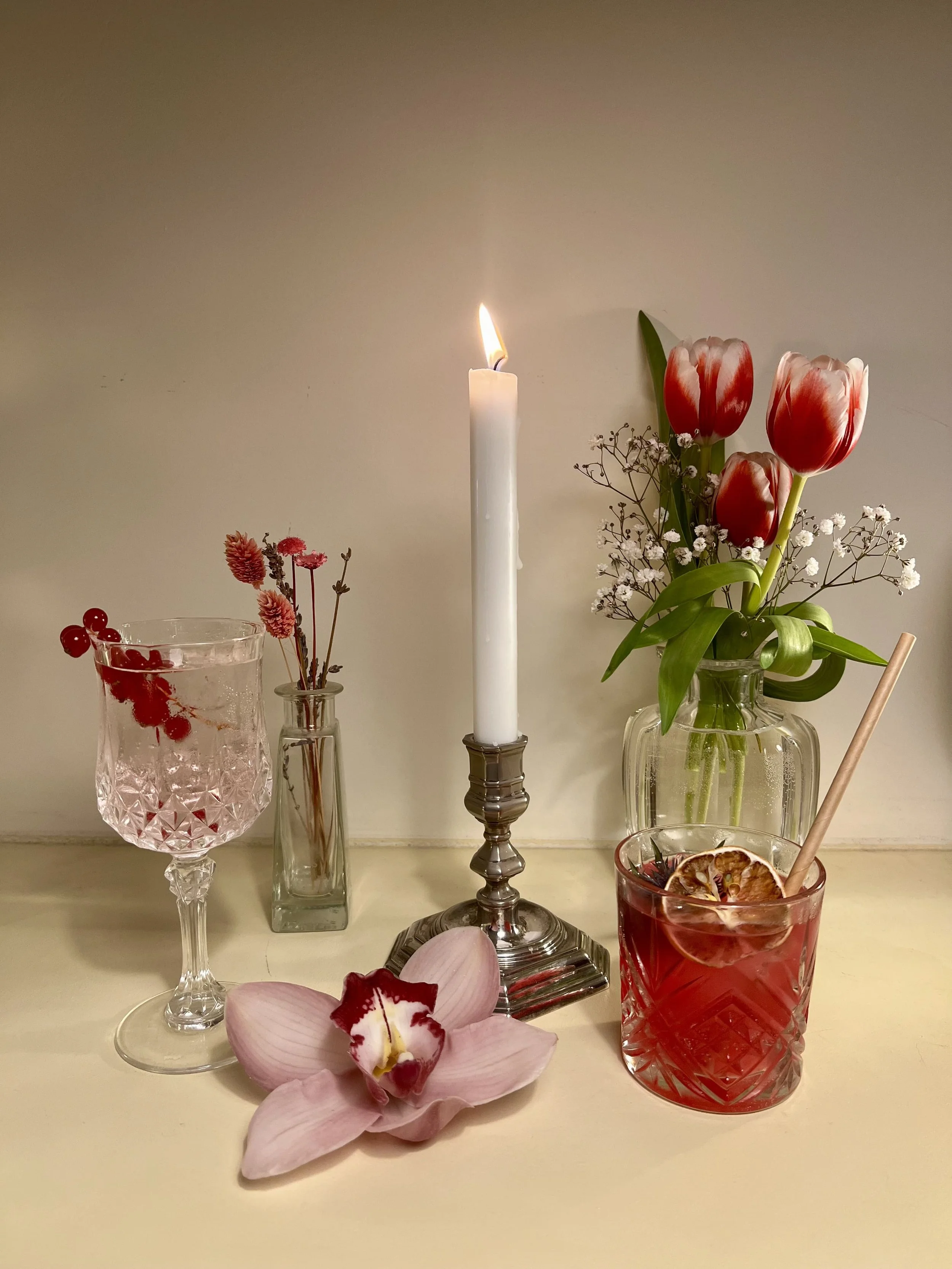 Trois verres sur une table, un contenant un cocktail rouge avec une tranche d'agrume, un autre avec des baies rouges et de l'eau gazeuse. Il y a une fleur orchidée rose devant, un vase avec des tulipes rouges et blanches à côté, et une bougie blanche