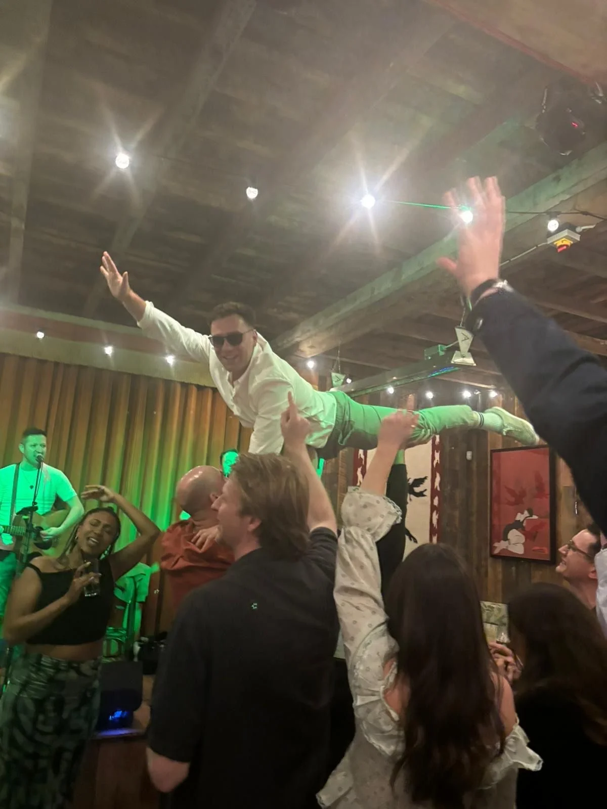 People at a wedding party, some dancing and one man being held horizontally above the crowd, smiling and wearing sunglasses, with a band performing in the background.