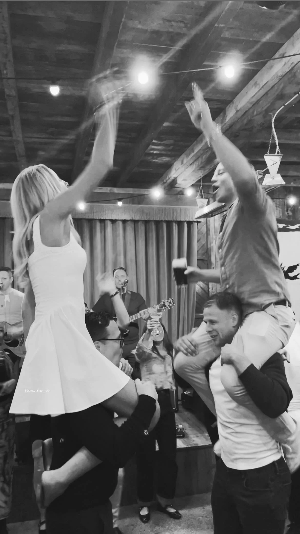People celebrating at a lively party, with two women on the shoulders of men dancing and high-fiving, in a rustic venue with string lights and a band playing in the background.