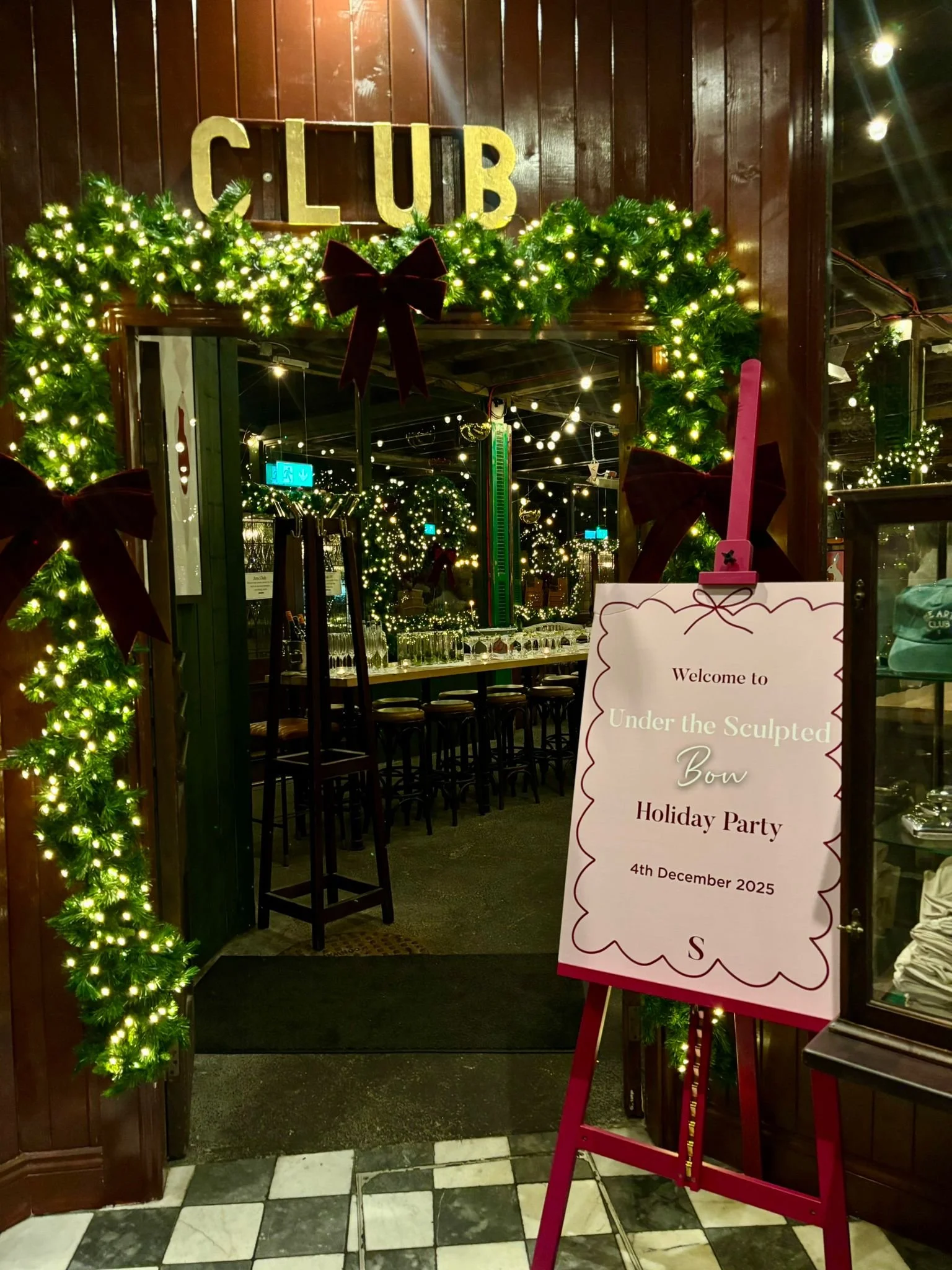Decorated entrance for a Christmas party with holiday greenery and lights. A sign reading 'Welcome to Under the Sculpted Bow Holiday Party, December 4, 2025'