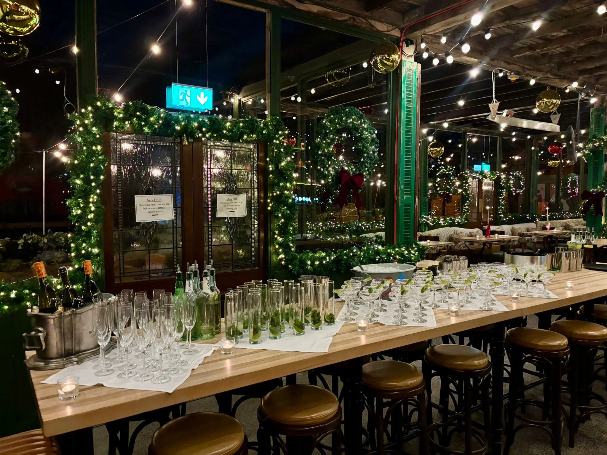 Festively decorated room interior with Christmas wreaths, string lights, and holiday ornaments. A long table is set with glasses, bottles, cocktails and candles, prepared for a celebration.