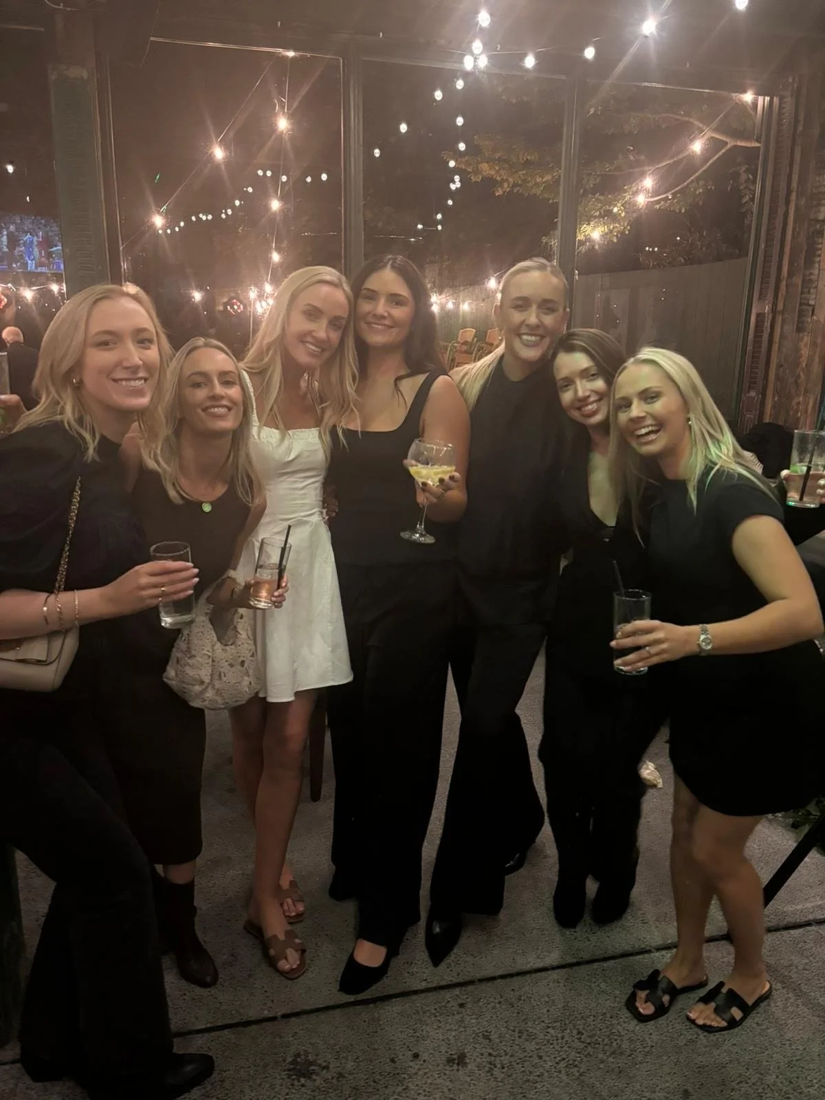 A group of seven women smiling and posing together at a social event, holding drinks, with warm lighting and decorative string lights in the background.