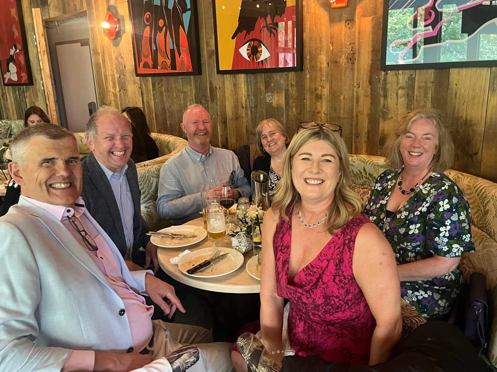Group of six adults sitting around a dinner table in a restaurant, smiling. The table has used plates and glasses, and the restaurant has wood-paneled walls and colorful artwork.