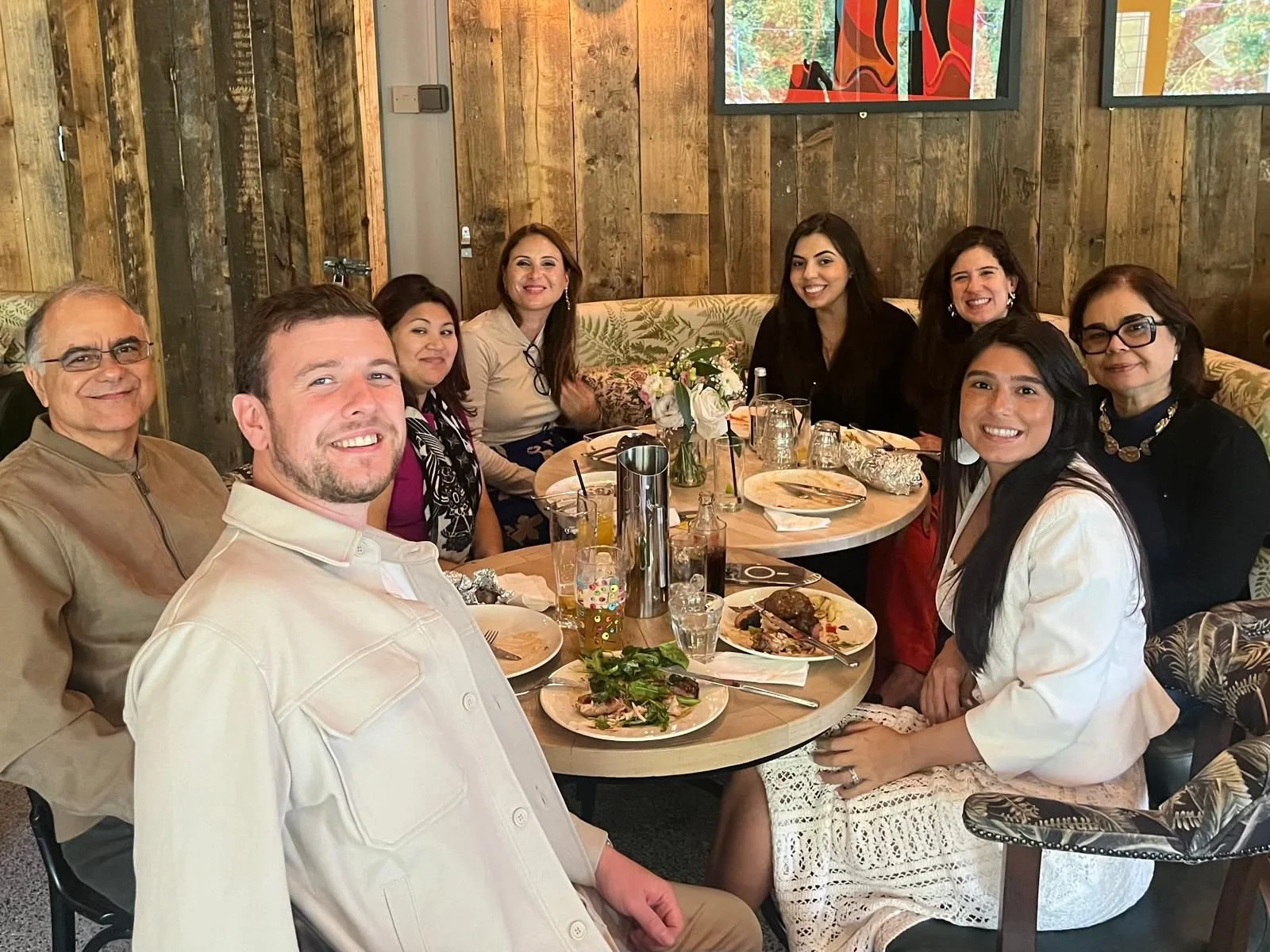 A group of nine people sitting around a round table in a restaurant, smiling and enjoying a meal, with plates of food and drinks on the table.