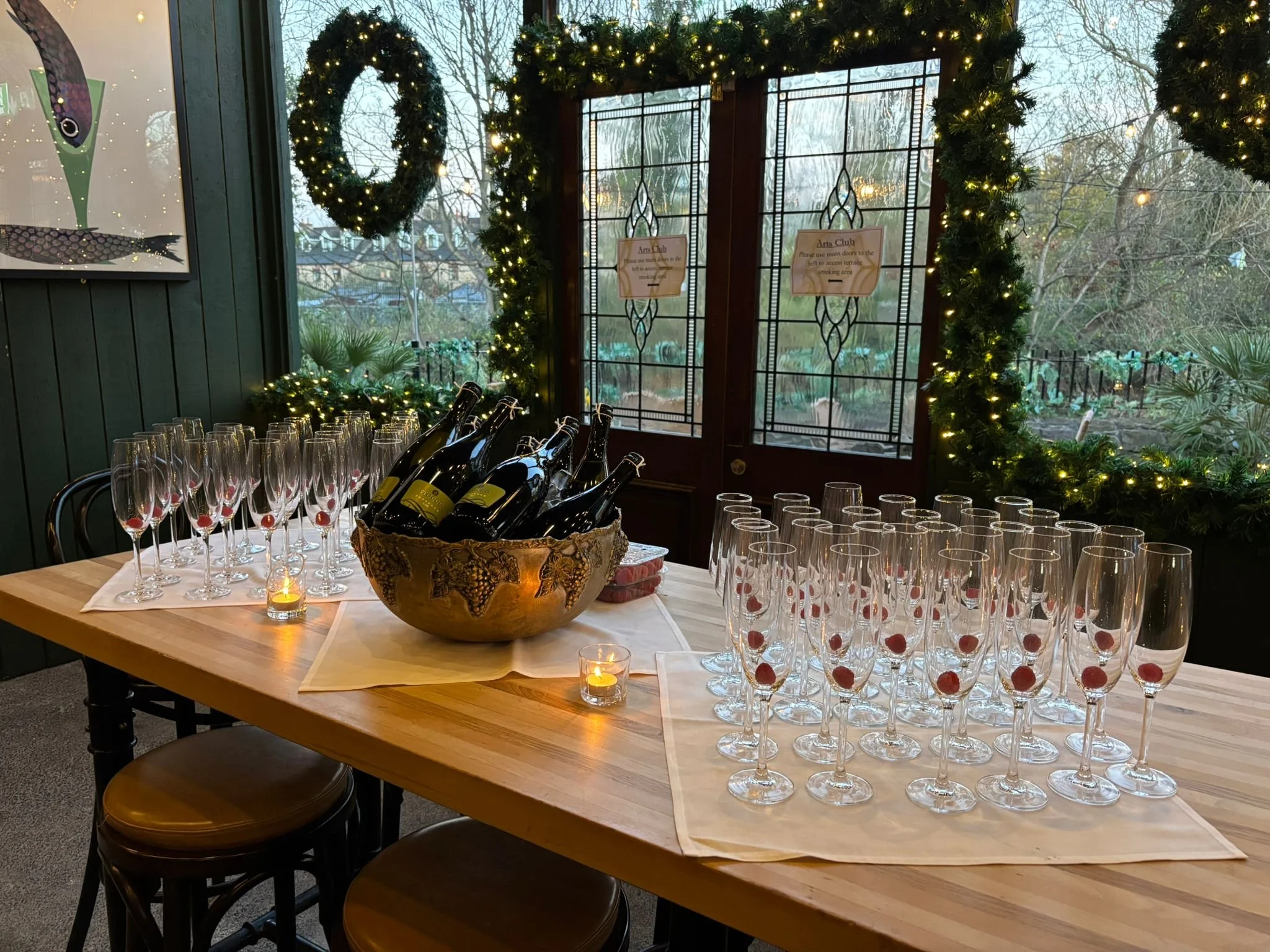 A wooden table with champagne glasses in a decorated event space. The glasses have raspberries inside and a silver ice bucket next to th.  In the background are windows and doors decorated with green garlands and lights, with a view of trees outside.