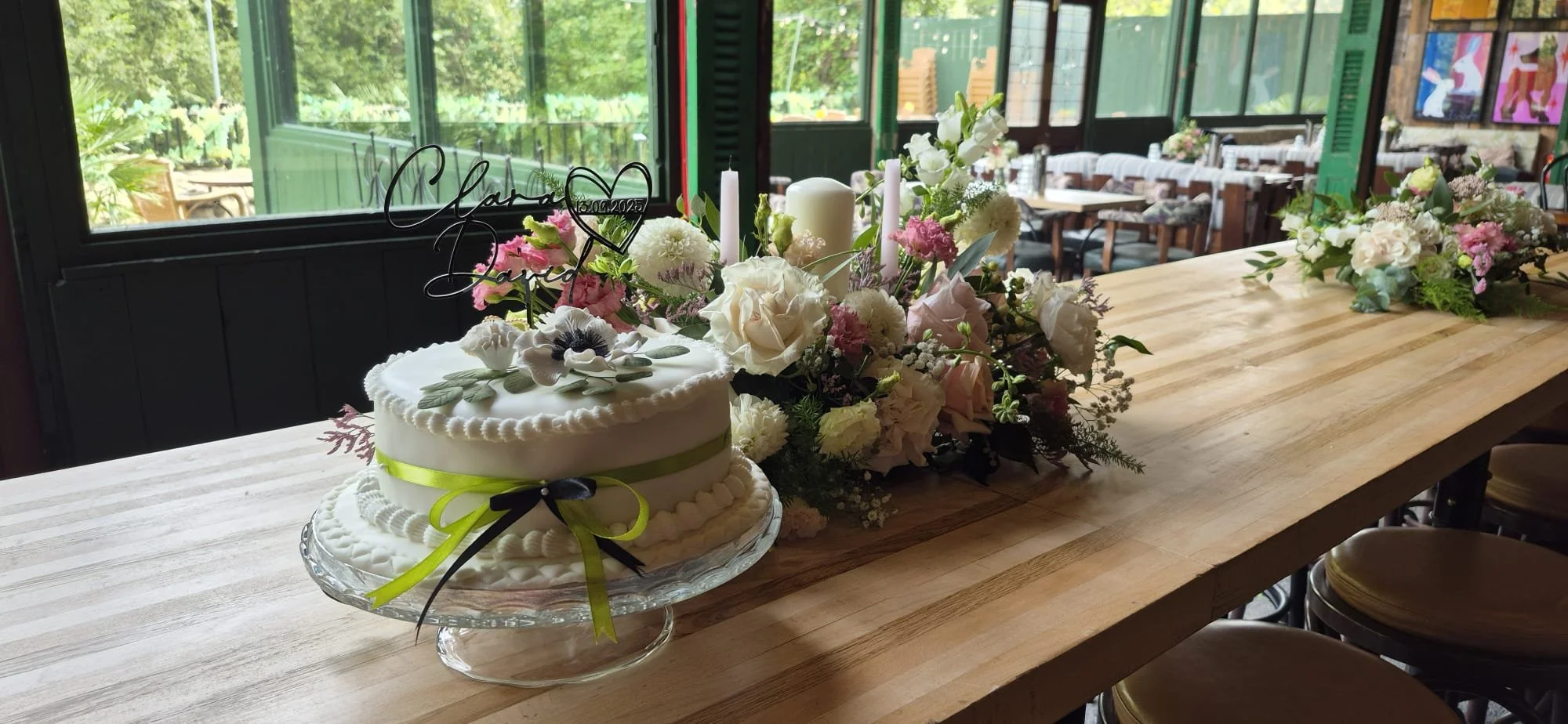Wedding cake decorated with white icing, flowers, and a green ribbon, placed on a glass stand on a wooden table surrounded by floral arrangements.