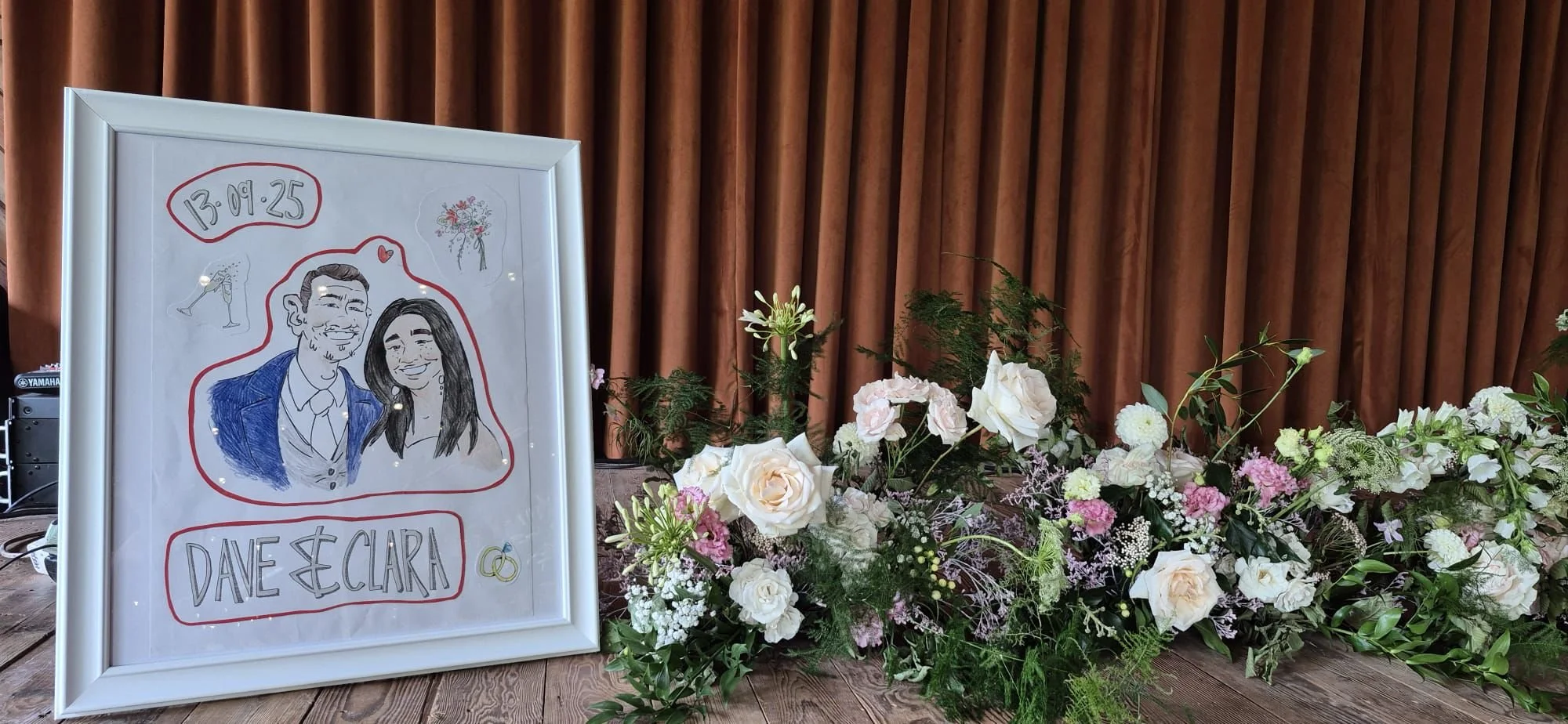 White framed drawing of a couple with wedding date, surrounded by floral arrangements on a wooden surface against a brown curtain background.