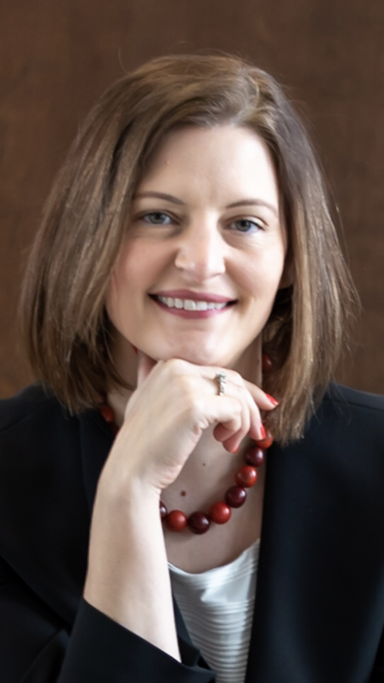 A woman with shoulder-length brown hair smiling, wearing a black blazer, white top, red beaded necklace, and a ring, with her hand resting on her chin.