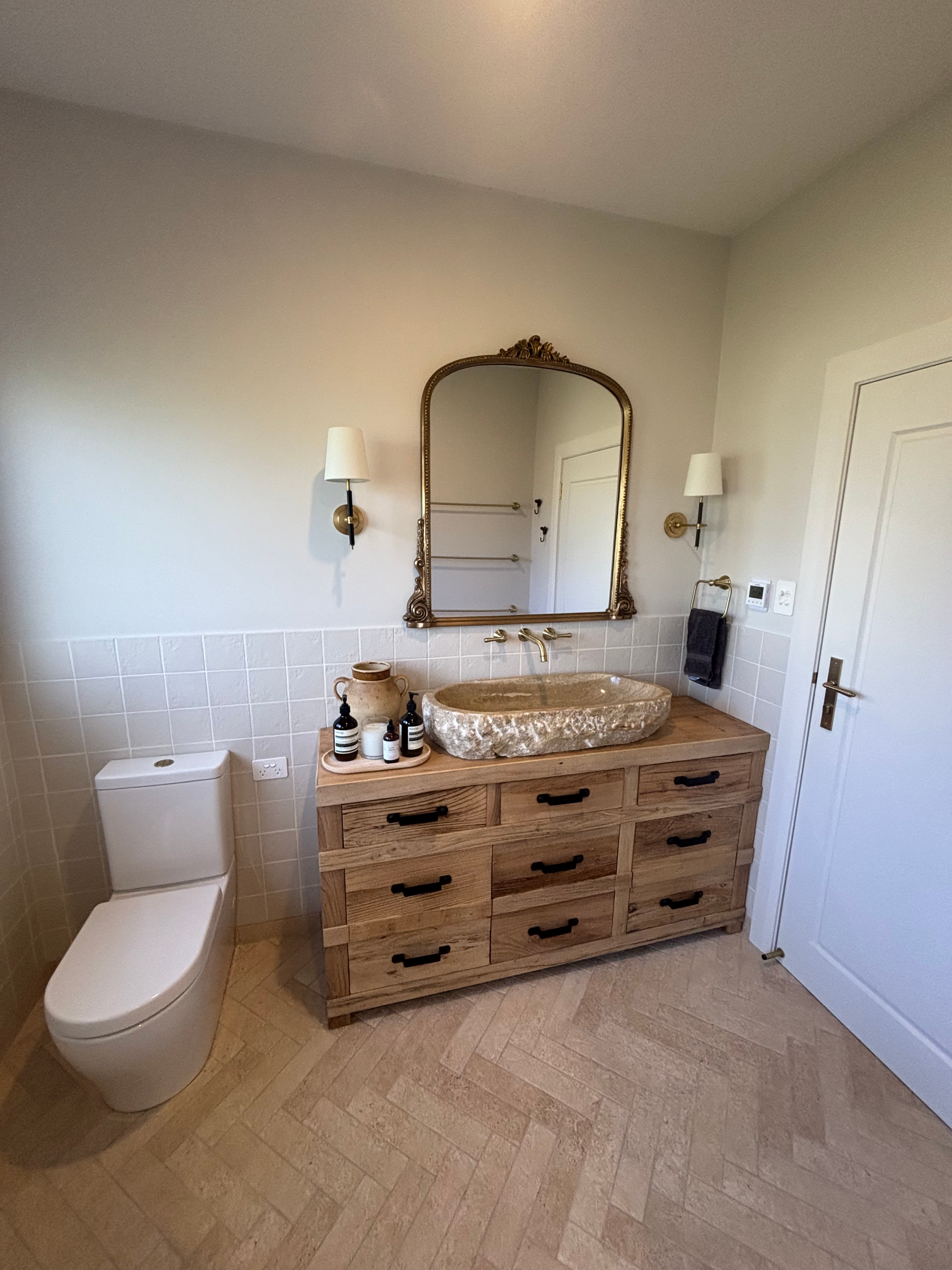 A bathroom renovation in Bowral, NSW in corner with a wooden vanity, oval sink, mirror, wall-mounted lights, toilet, and decorated items.