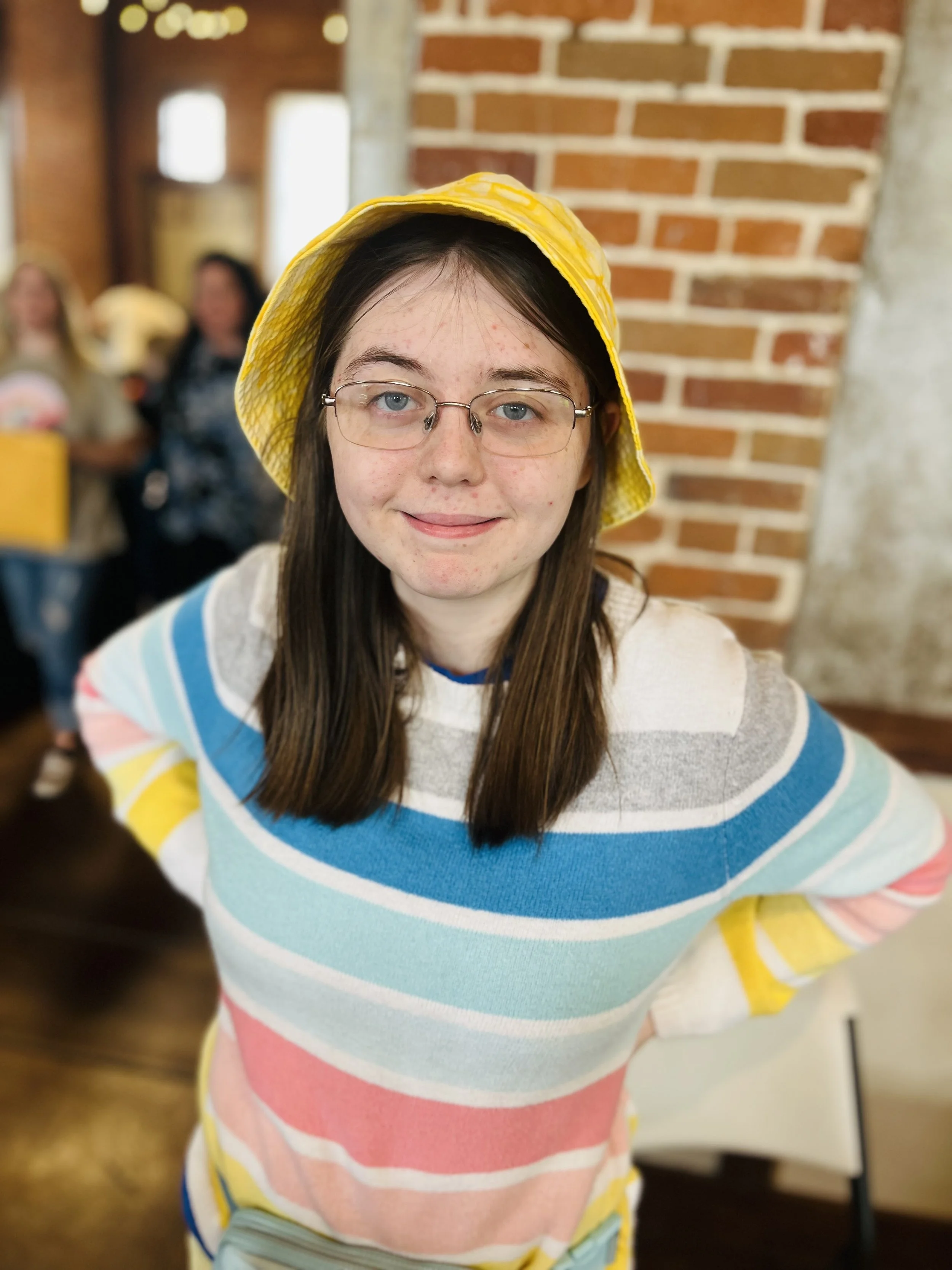 A young woman with glasses and long brown hair wearing a yellow hat and a colorful striped sweater, smiling with her hands on her hips, in an indoor setting with brick walls and blurred people in the background.