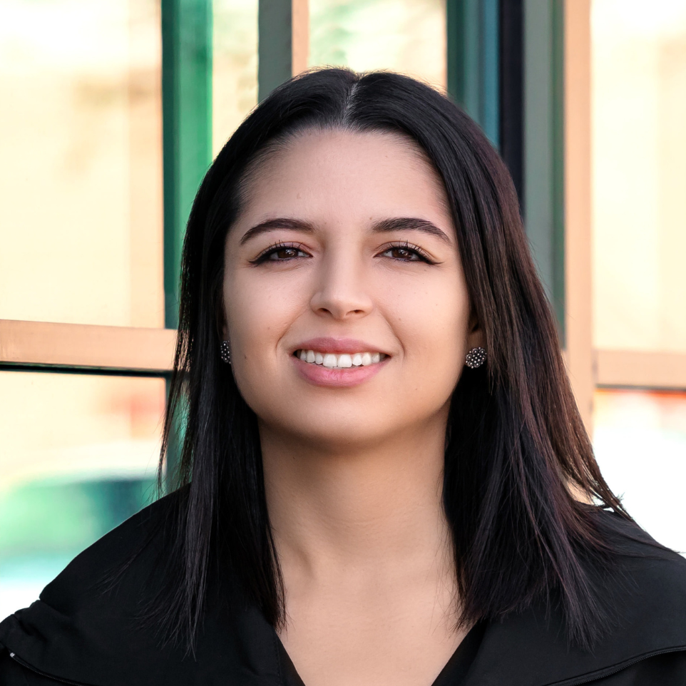 Young woman with shoulder-length dark hair, smiling, in front of a colorful fence background.