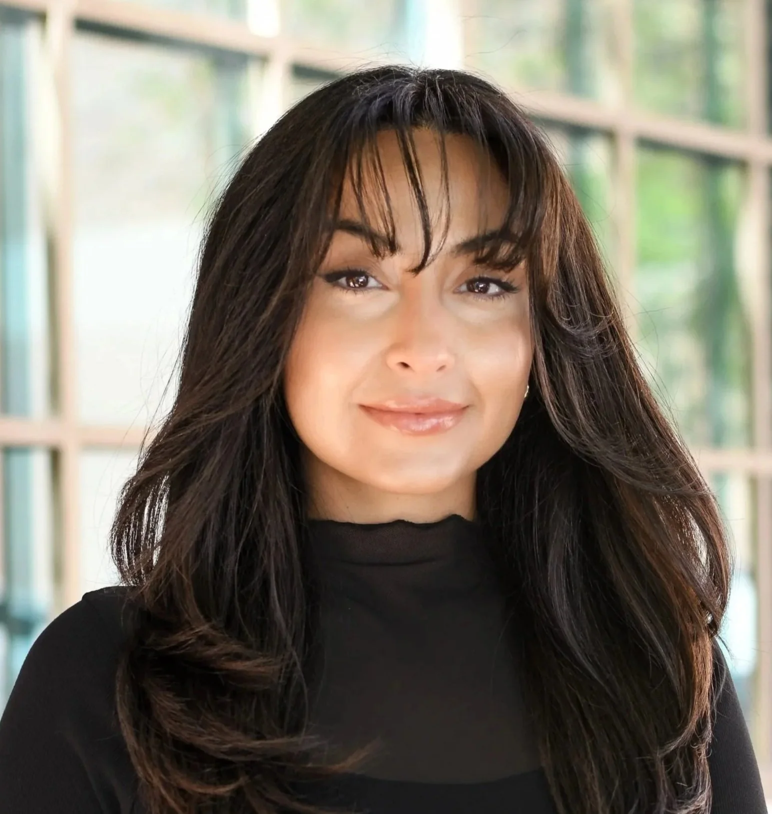 A woman with long dark wavy hair standing outdoors in front of a brick wall and a window, smiling at the camera.