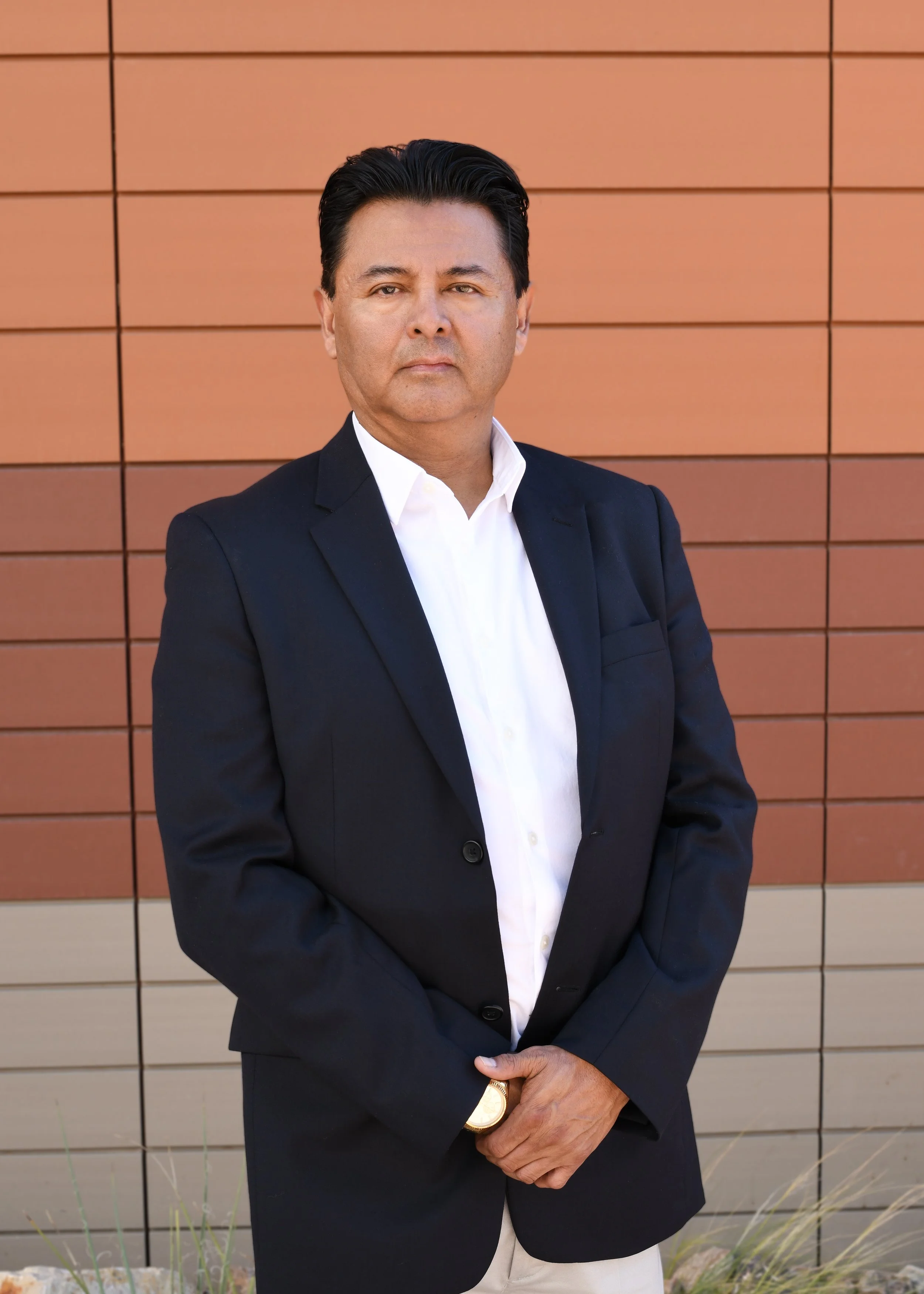 Portrait of a man with dark hair, wearing a white collared shirt, standing outdoors near a building with glass windows and brick wall in the background.