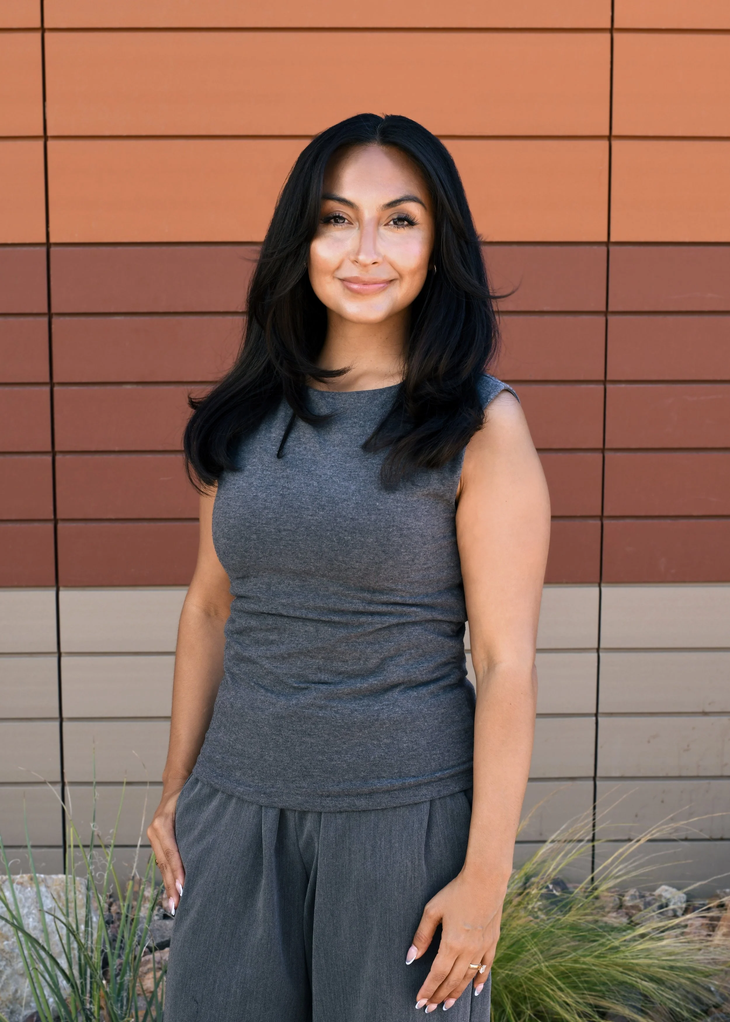 A woman with long dark wavy hair standing outdoors in front of a brick wall and a window, smiling at the camera.