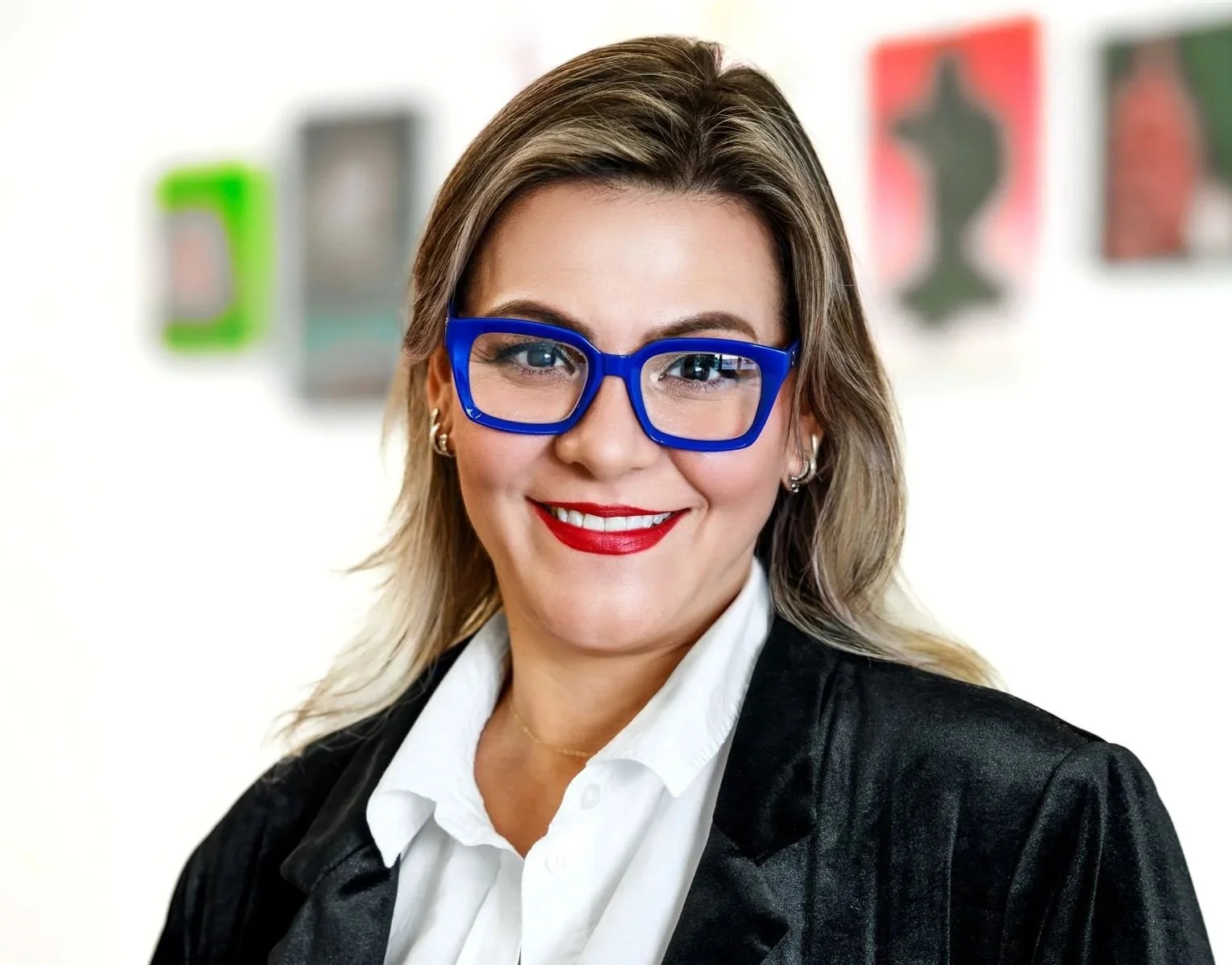 Woman with blonde hair wearing large blue glasses, red lipstick, white shirt, and black blazer, smiling in front of a white wall with colorful abstract art.