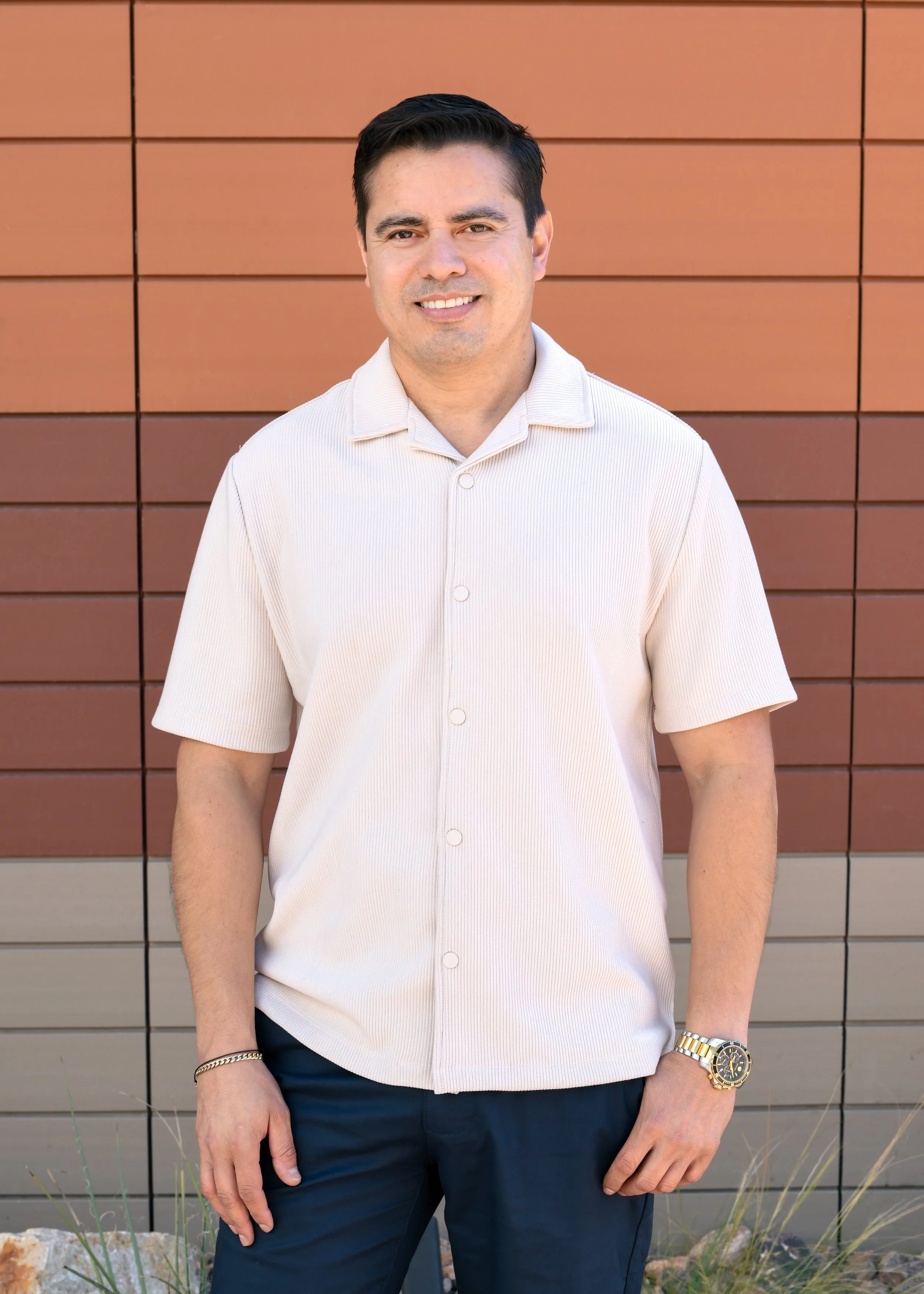 A man with short dark hair wearing a black polo shirt standing indoors with a blurred background that includes orange and white elements.
