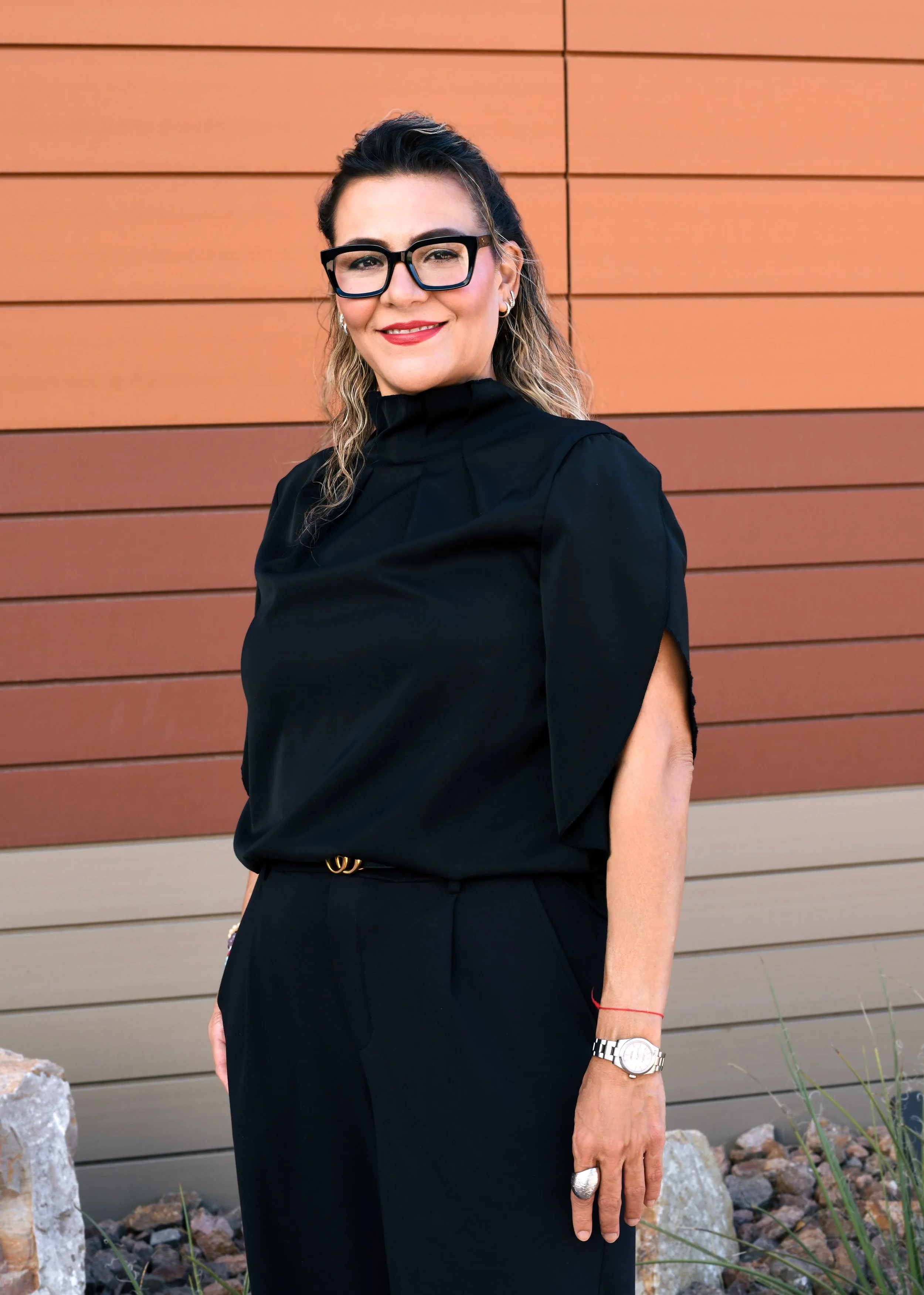 Woman with blonde hair wearing large blue glasses, red lipstick, white shirt, and black blazer, smiling in front of a white wall with colorful abstract art.