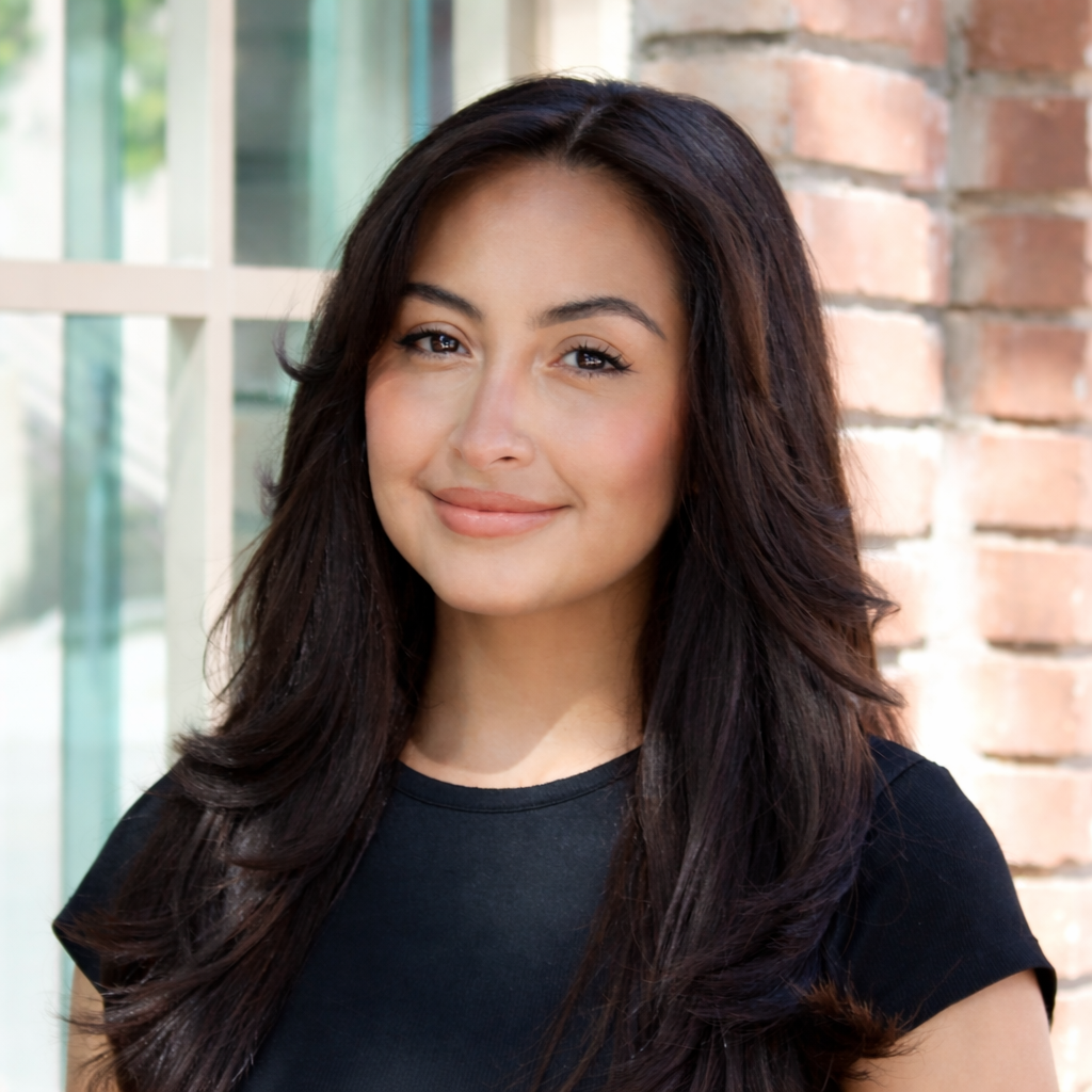 A woman with long dark wavy hair standing outdoors in front of a brick wall and a window, smiling at the camera.