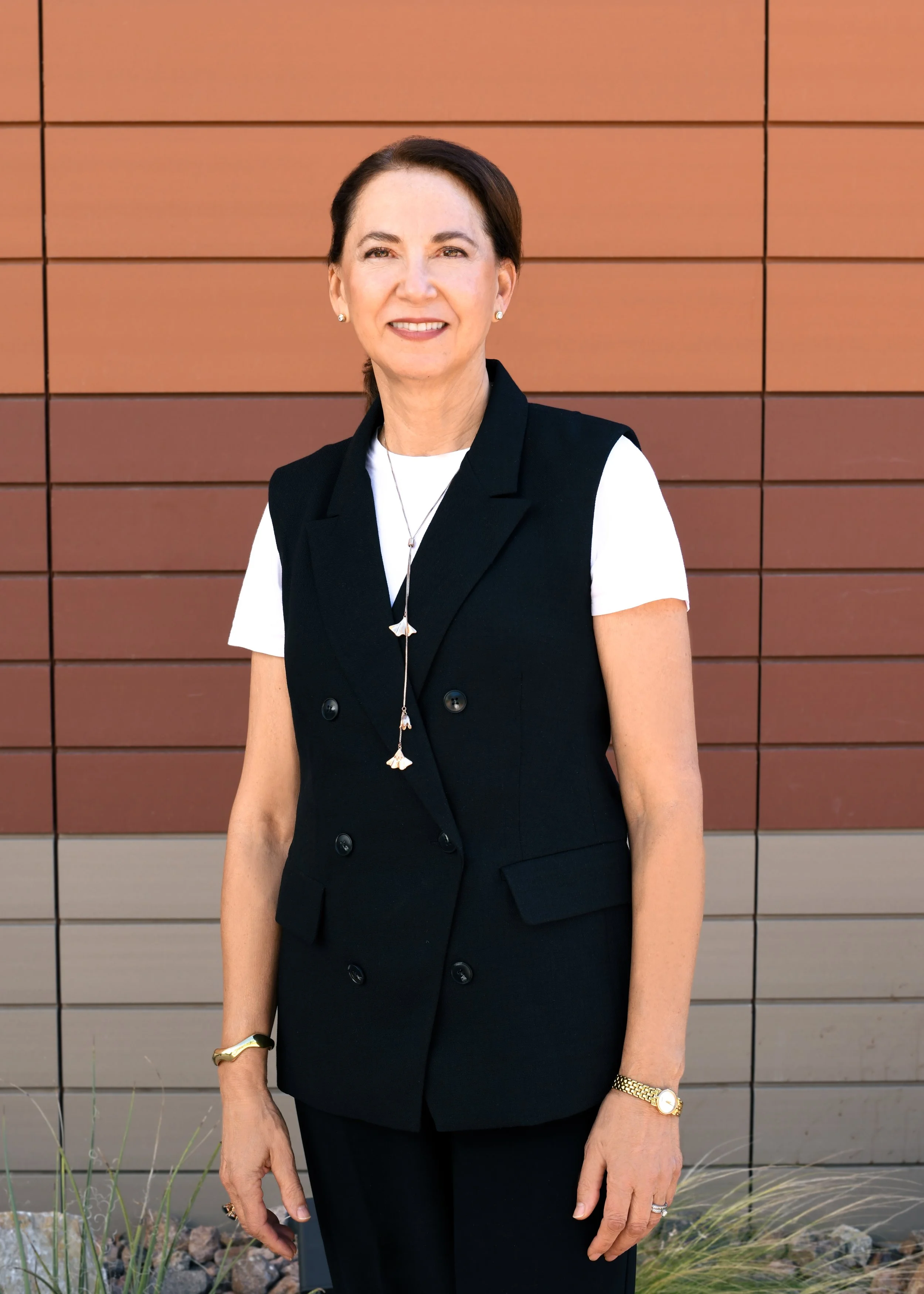 A woman with brown hair smiling, wearing a white blouse with green and black leaf patterns, gold and silver jewelry, standing outdoors near a building with windows and brick wall.