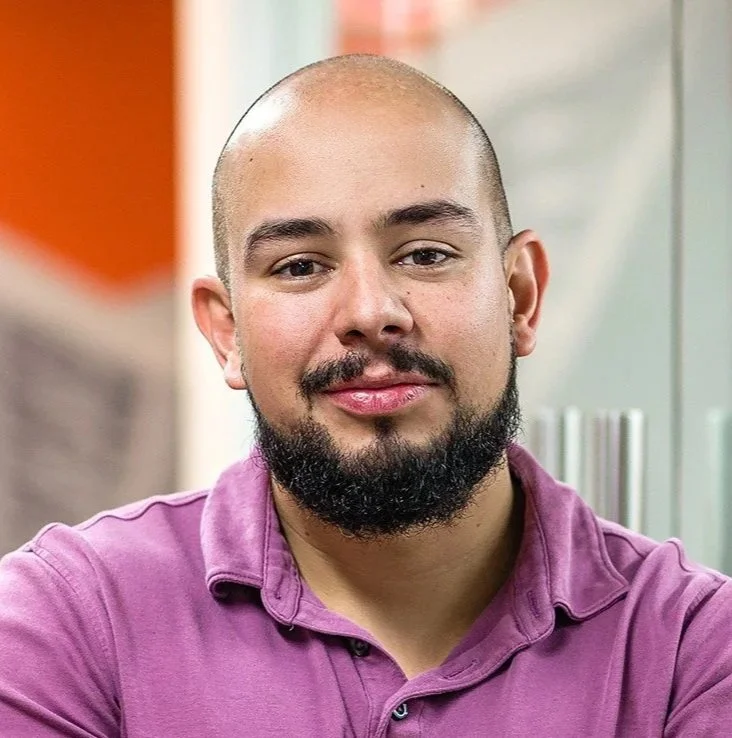 A young man with a shaved head and beard, wearing a purple shirt, smiling and sitting indoors.