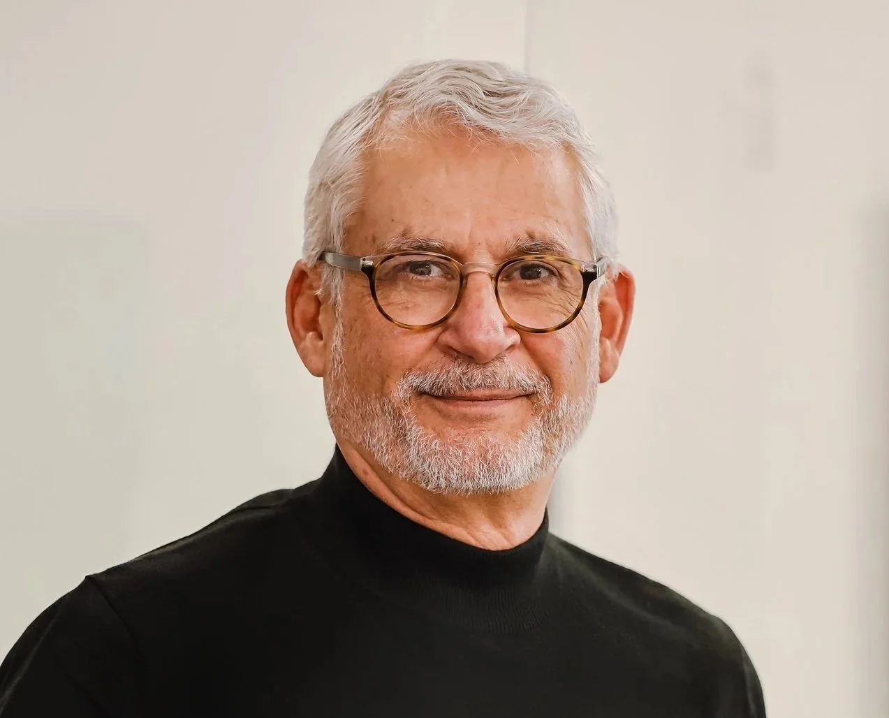 Older man with gray hair, glasses, and a beard, wearing a black turtleneck sweater, smiling at the camera indoors.