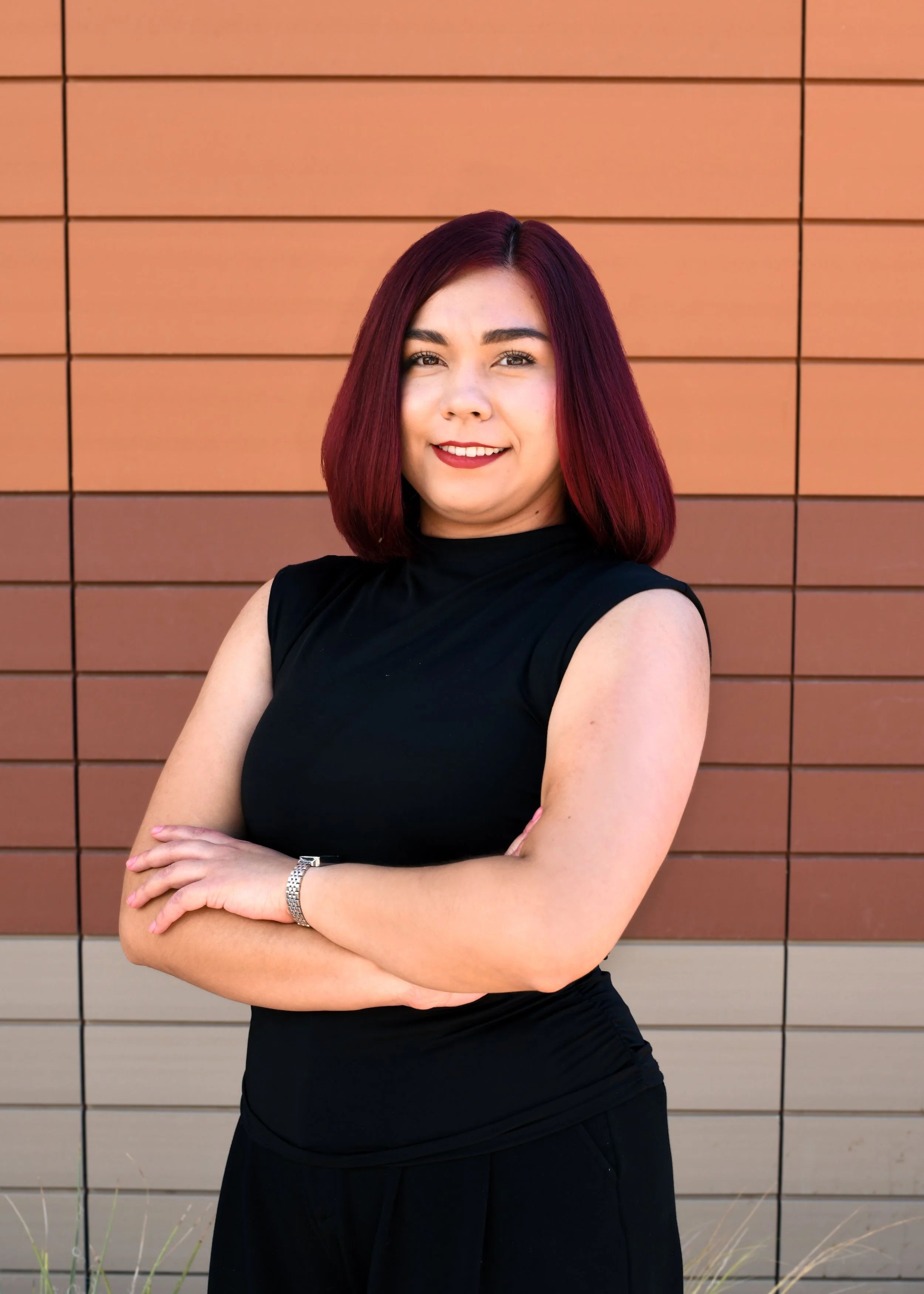 A woman with shoulder-length brown hair, wearing red lipstick and a black top, smiling outdoors with a blurred cityscape and blue sky in the background.