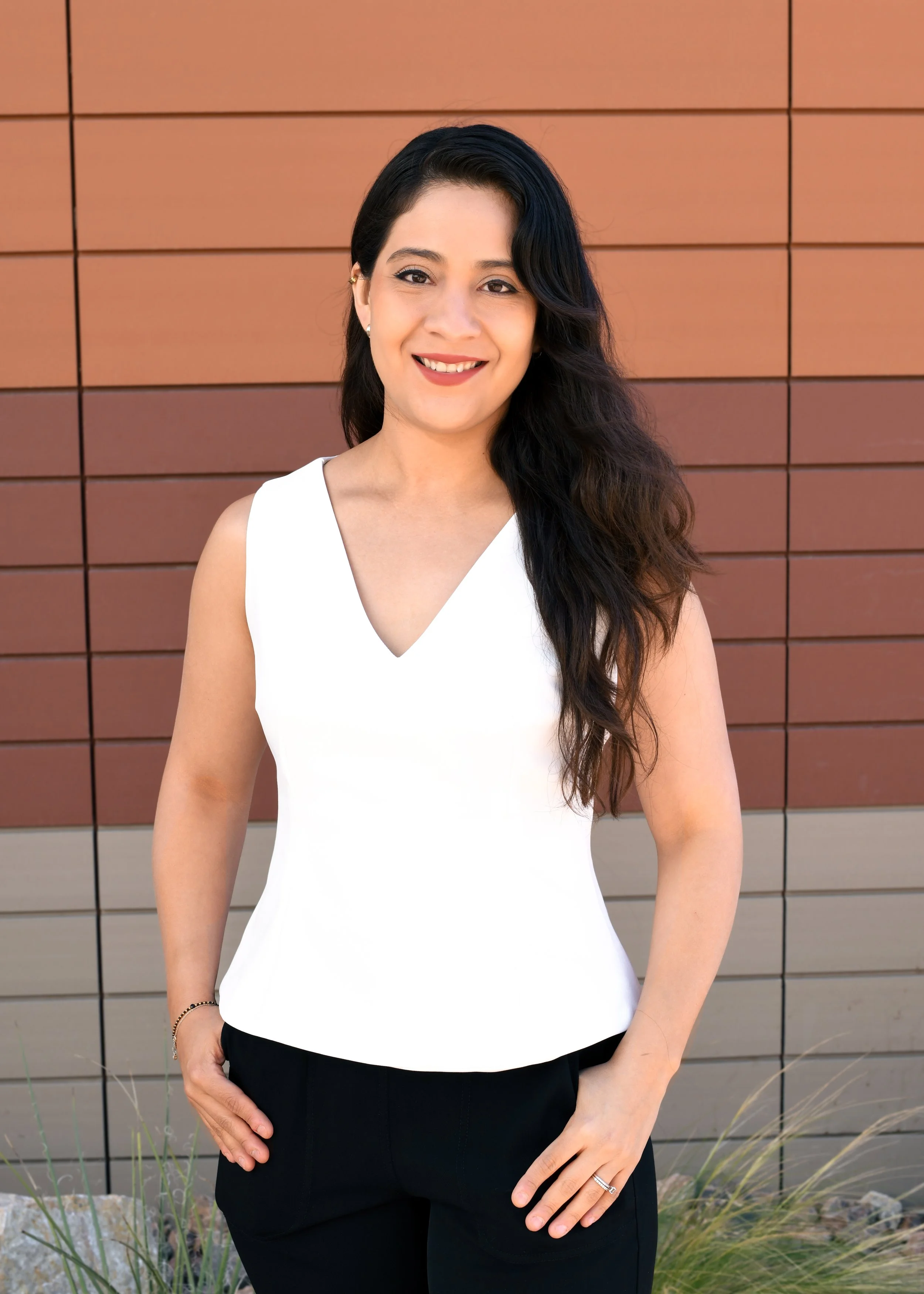 A young woman with long wavy dark brown hair, wearing a black and white checkered sleeveless top, smiling outdoors near a brick wall and window.
