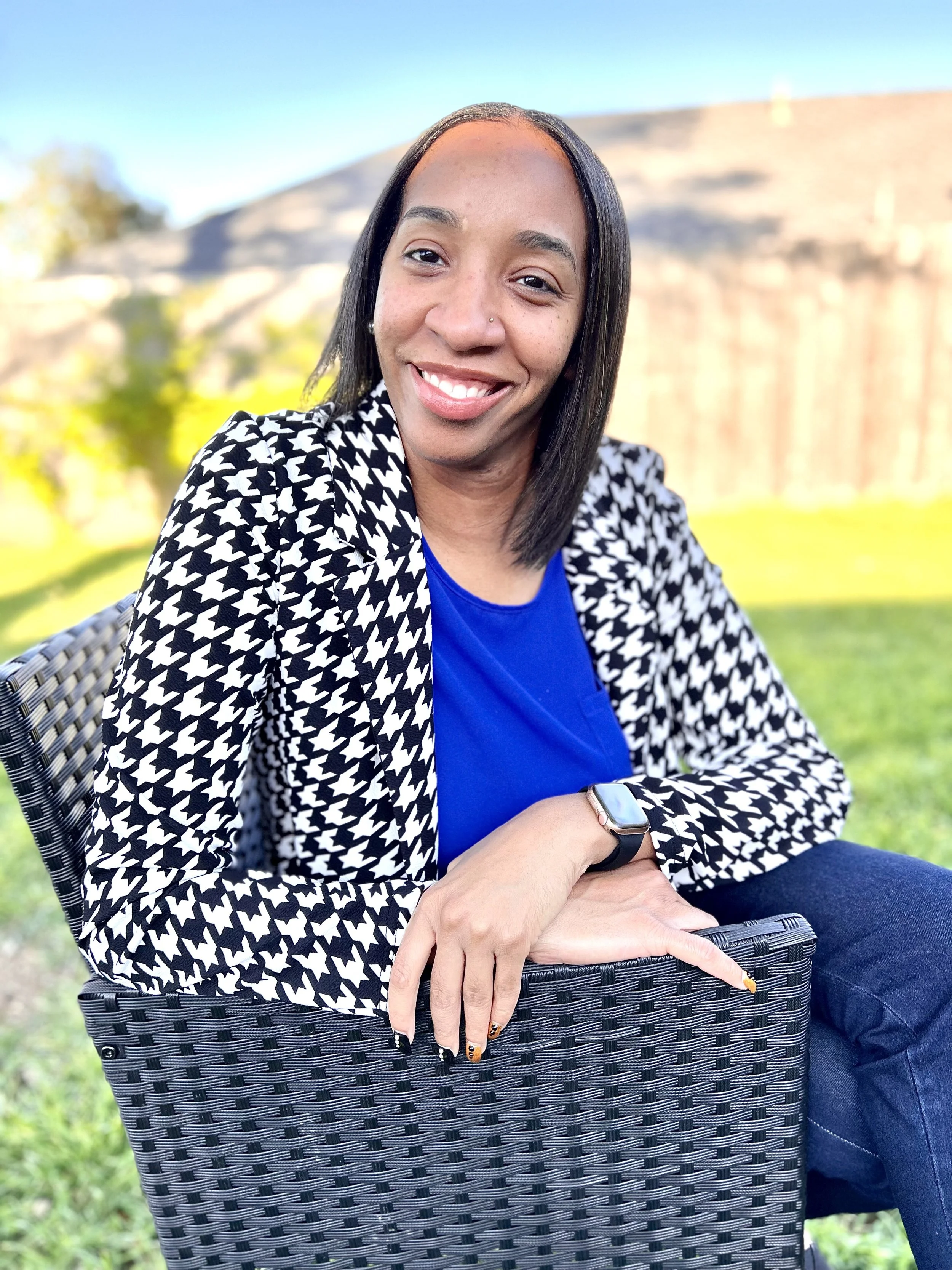 Smiling woman in a houndstooth blazer and blue top sitting outdoors on a chair at Tone Set Therapy