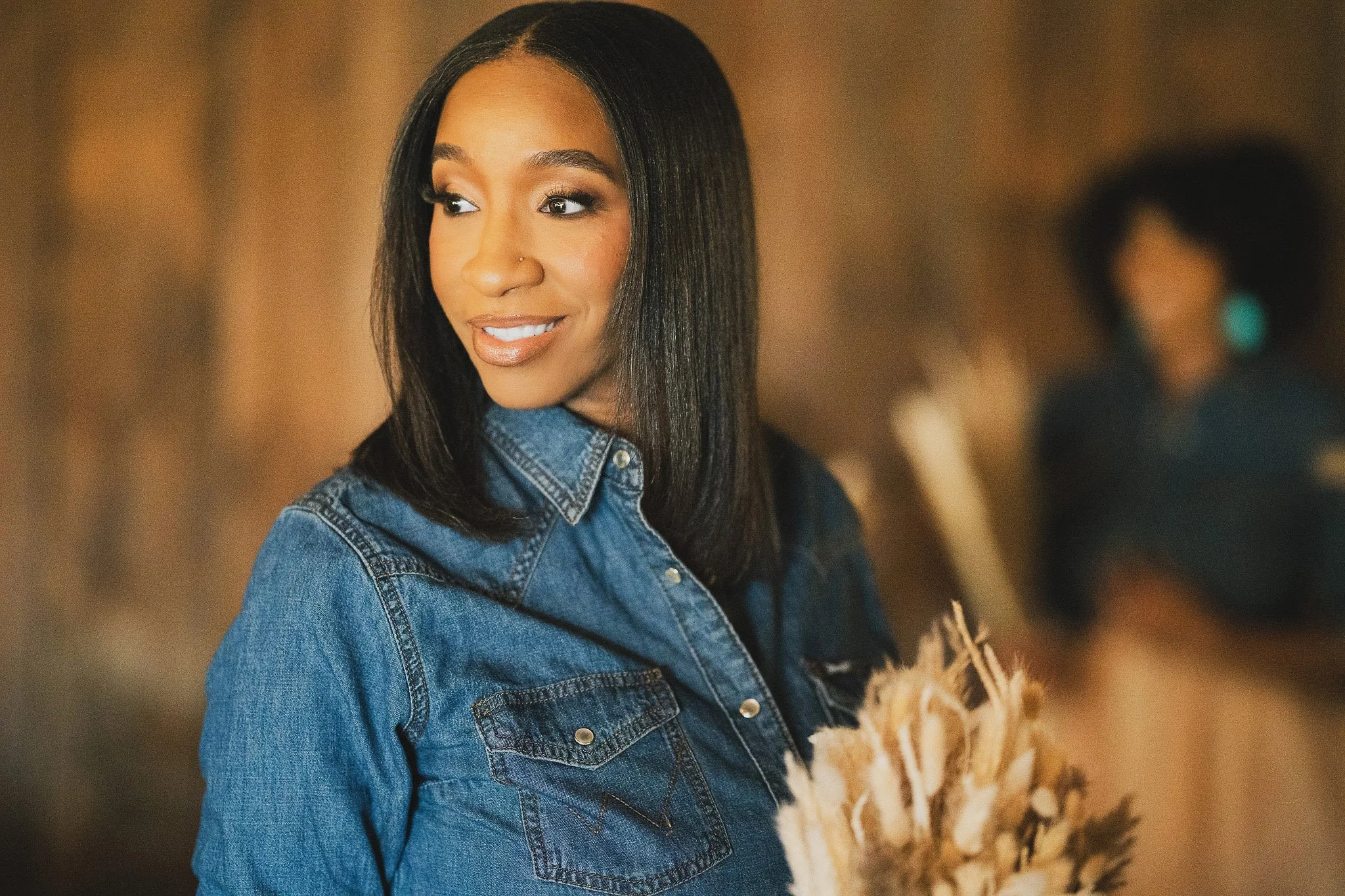 A woman with medium skin tone, straight black hair, and a nose piercing, wearing a blue denim jacket, looks to her left with a smile. Blurred background with other people.