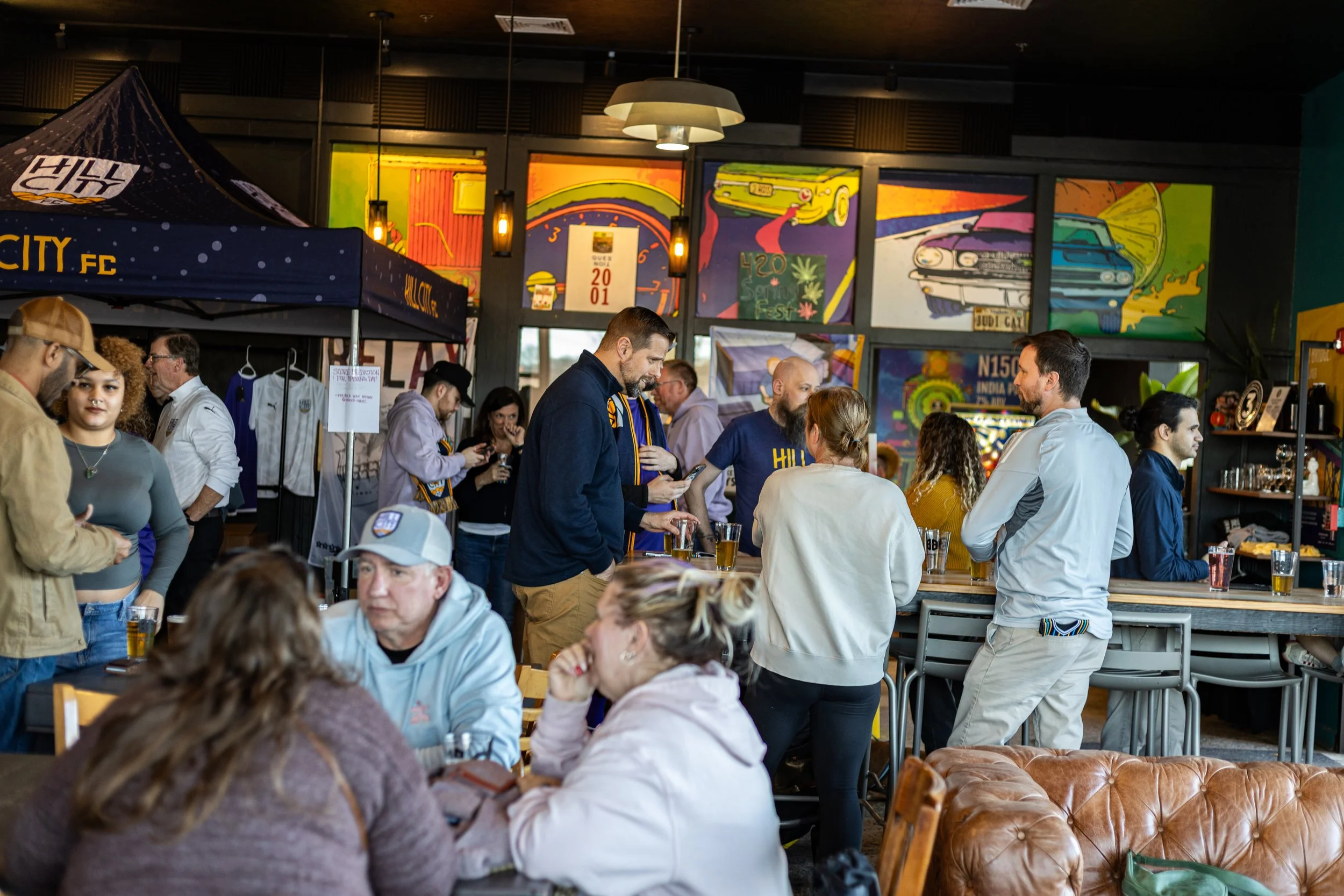 Indoor gathering at a bar or pub, with people socializing and ordering drinks, decorated with colorful automotive-themed graffiti paintings on the walls.
