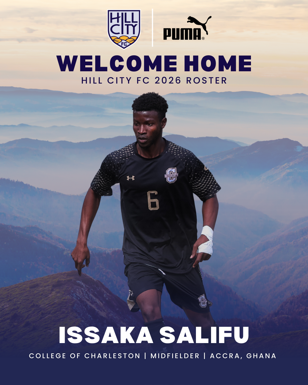A soccer player in a black Hill City FC jersey walking on a mountain trail with a scenic mountain landscape in the background.