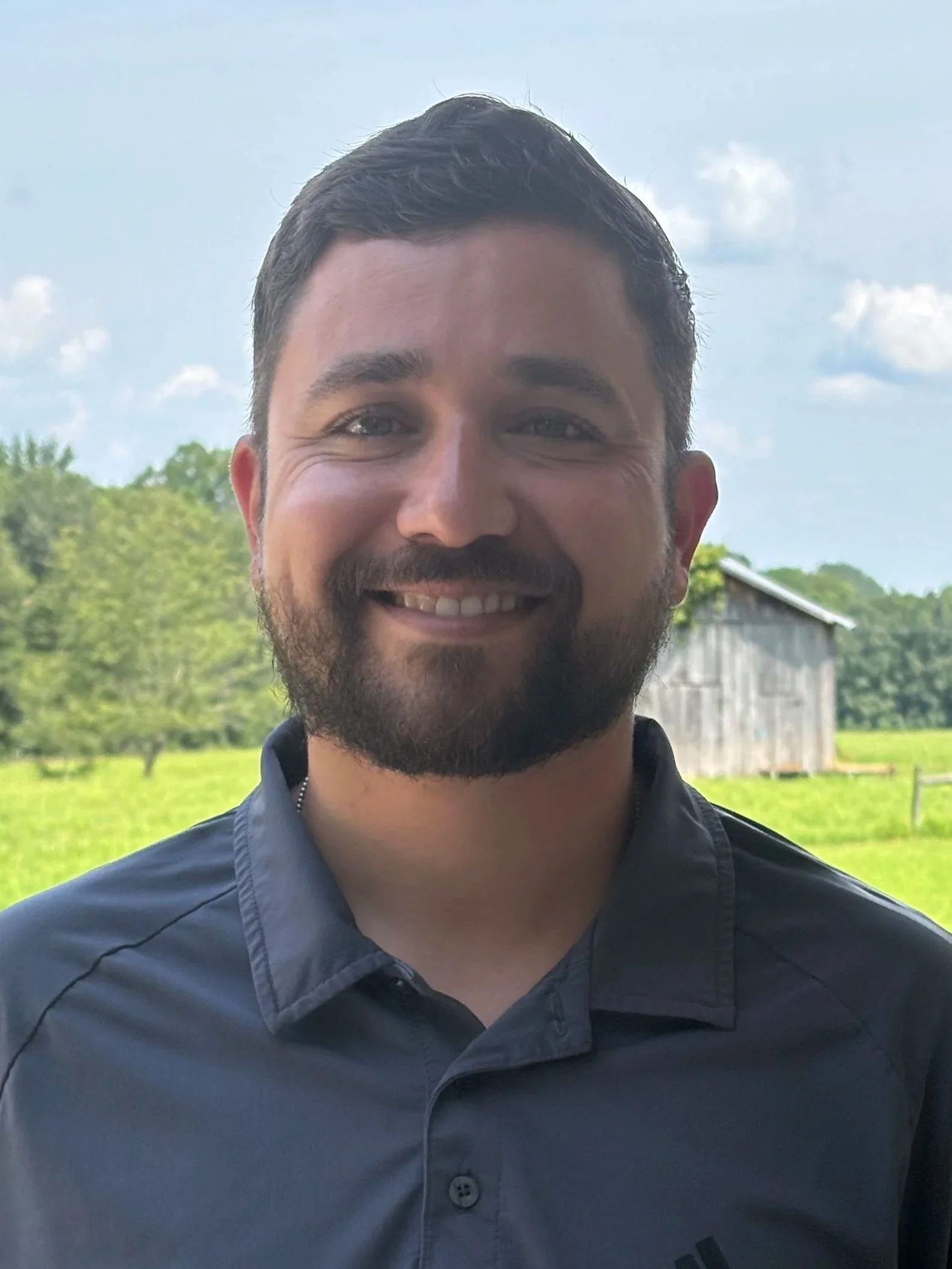 A smiling man with dark hair and a beard standing outdoors with a barn and green fields in the background on a partly cloudy day.