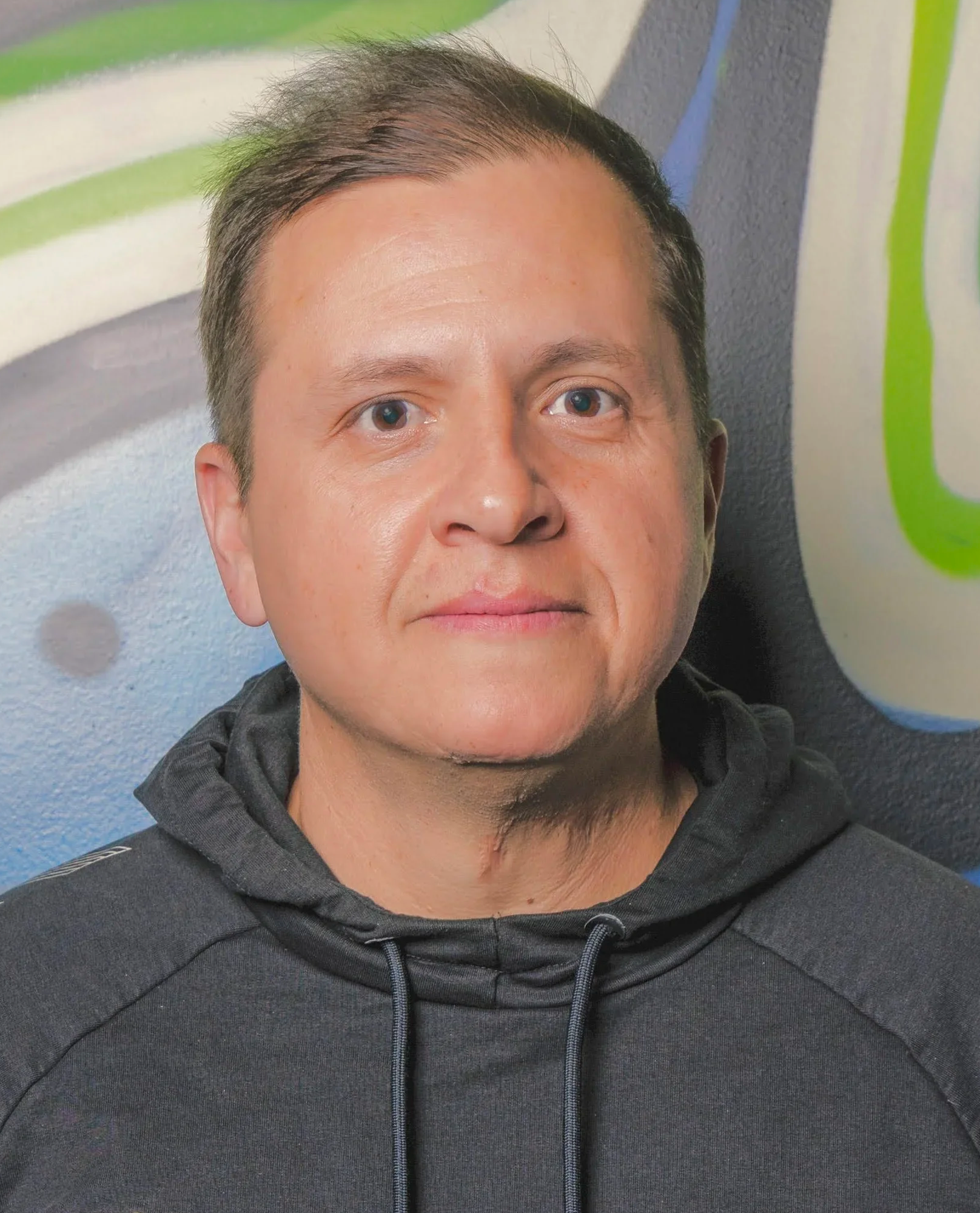 A man with short, dark hair and light skin, wearing a black hoodie, standing in front of a colorful graffiti wall.