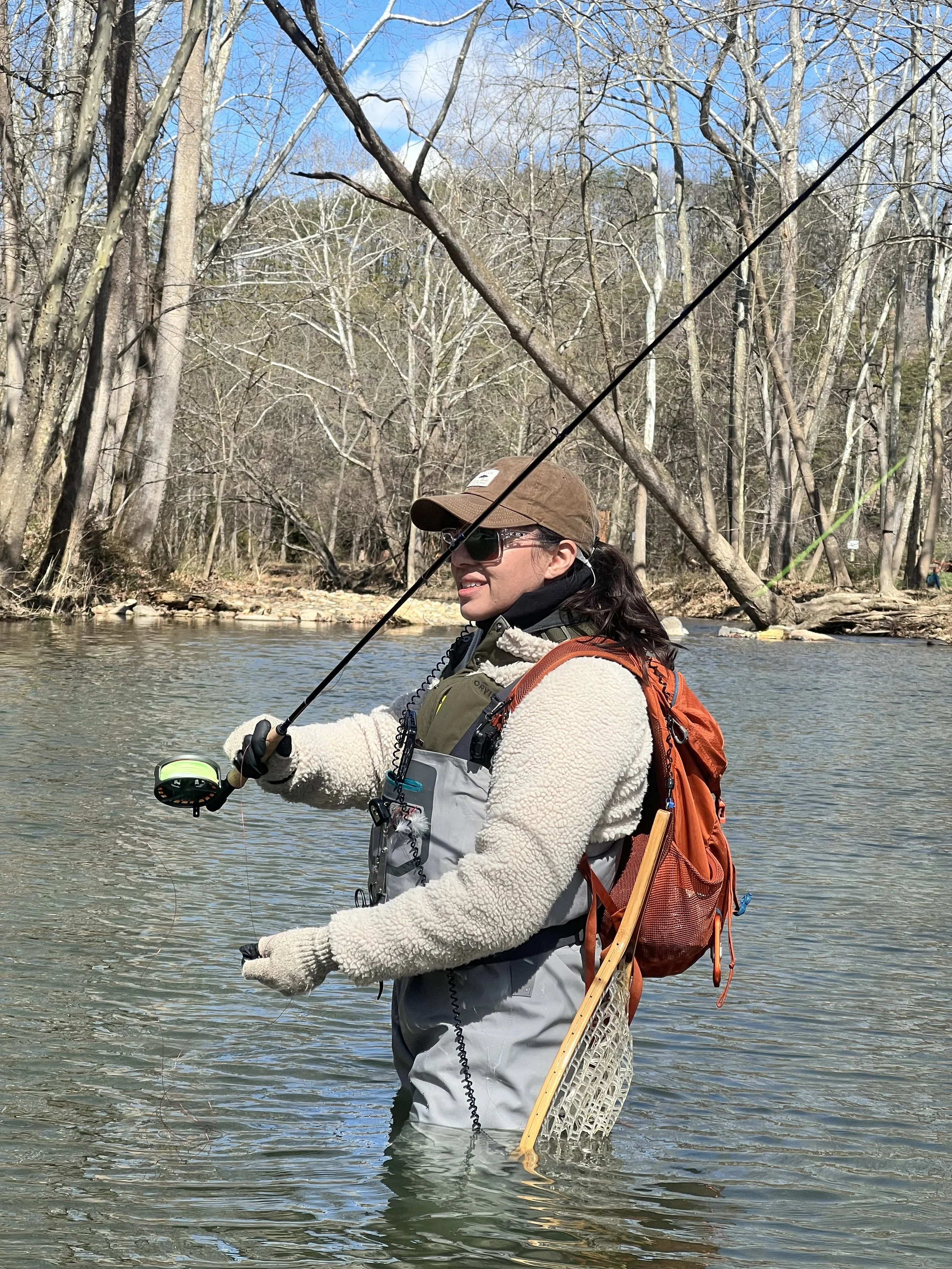 A woman fishing in a river, standing in waist-deep water, with trees and a clear sky in the background.