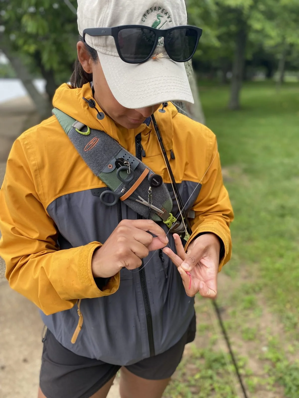 A woman in outdoor attire is preparing fishing gear, wearing a beige cap with sunglasses perched on top, a yellow and gray jacket, and a harness with fishing tools. She is holding a fishing hook and line in her hands, standing on a trail in a lush, green park or forested area.