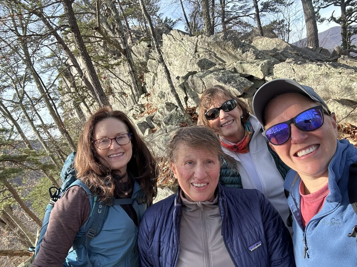 Four women hiking outdoors in a forested area with rocks, wearing outdoor gear and sunglasses, smiling at the camera.
